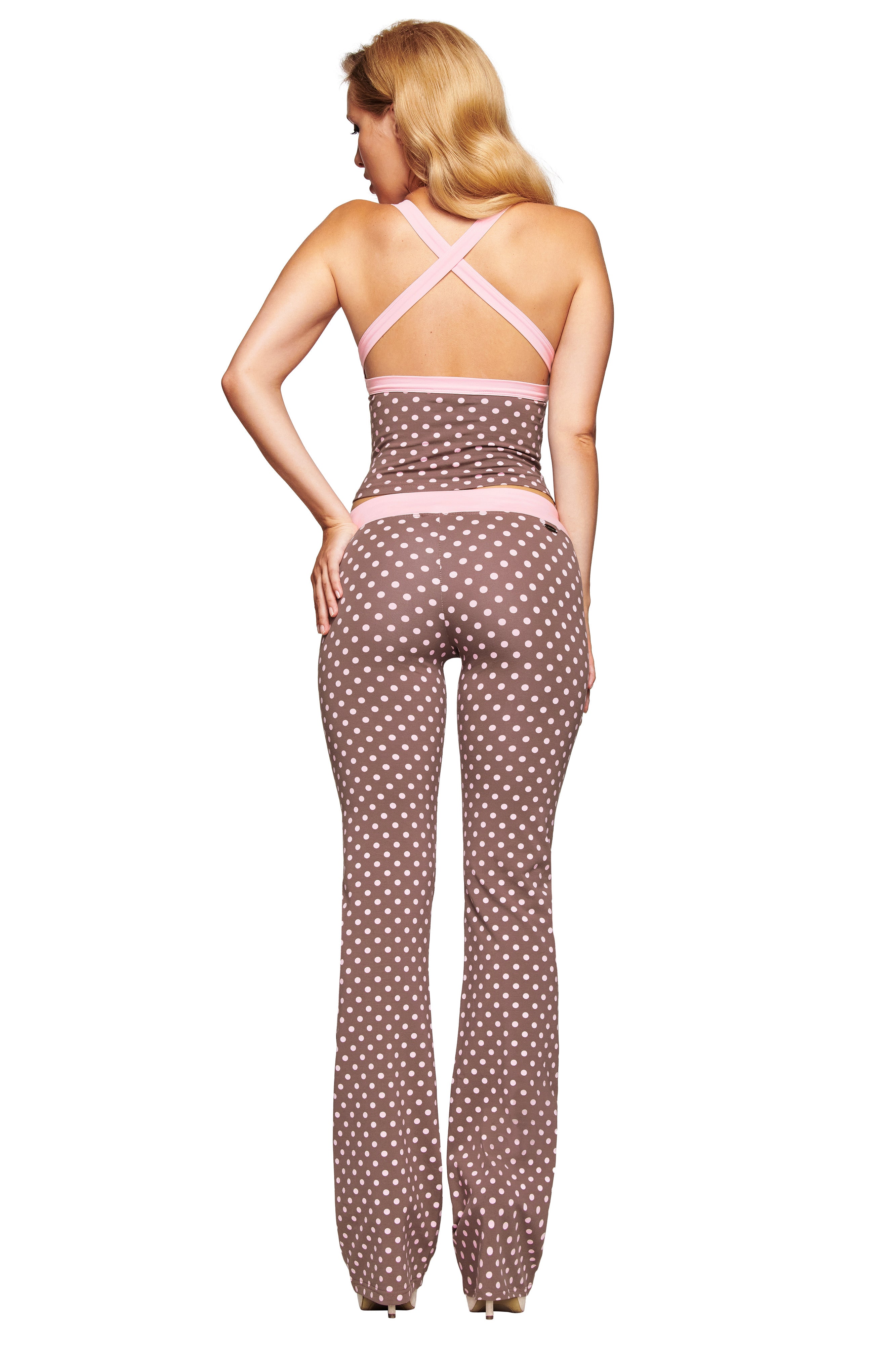 A woman with long blonde hair is seen from the back wearing the I.AM.GIA BLARE CROSSOVER PANT in Pink Chocolate, featuring a brown polka dot print, crisscross straps, flared legs, and a stylish mid-rise waist.