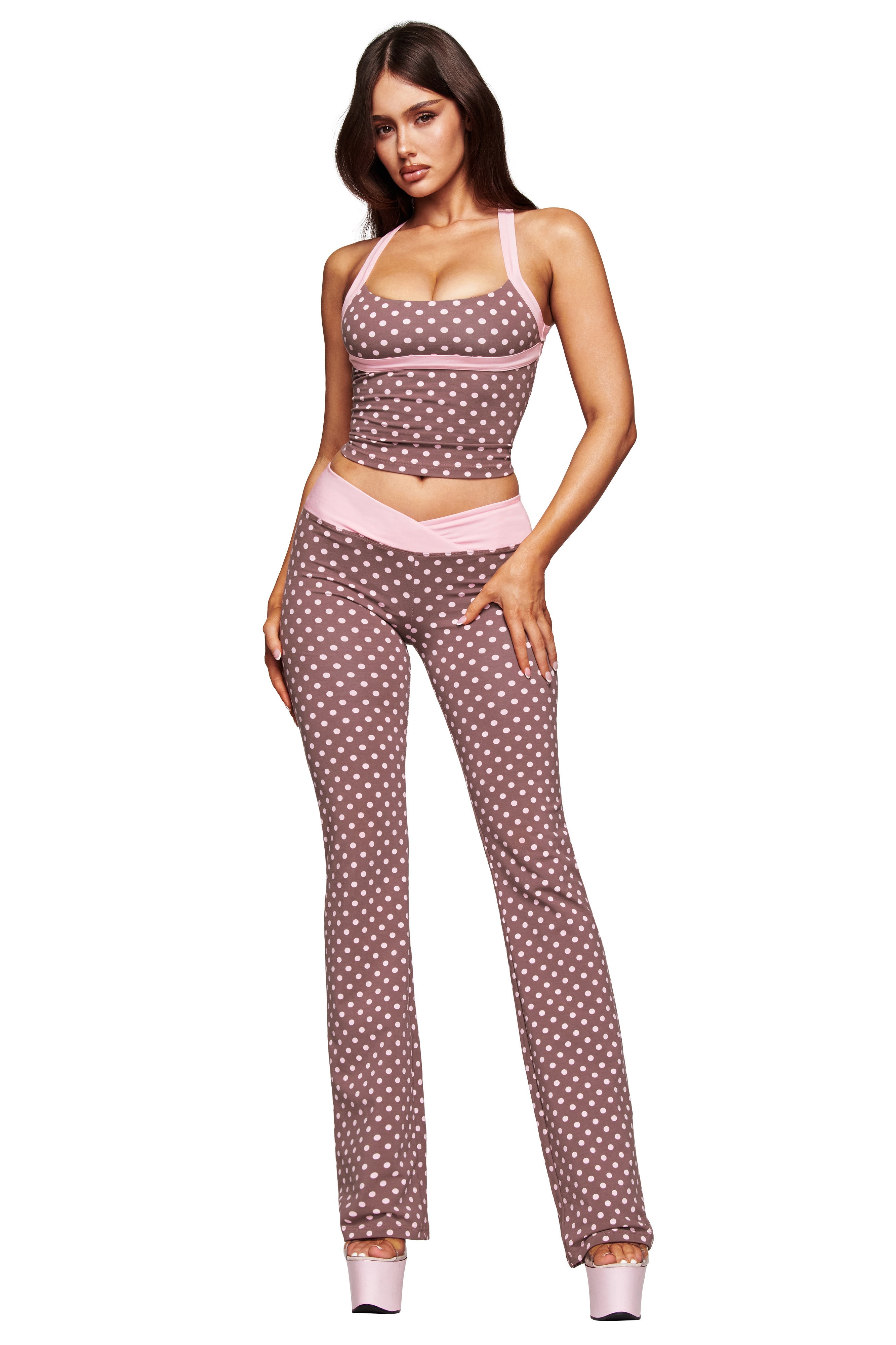 A woman models the I.AM.GIA BLARE CROSSOVER PANT in Pink Chocolate, featuring a polka dot print, light pink trim, and mid-rise fit, paired with a matching crop top and high platform heels against a white background.