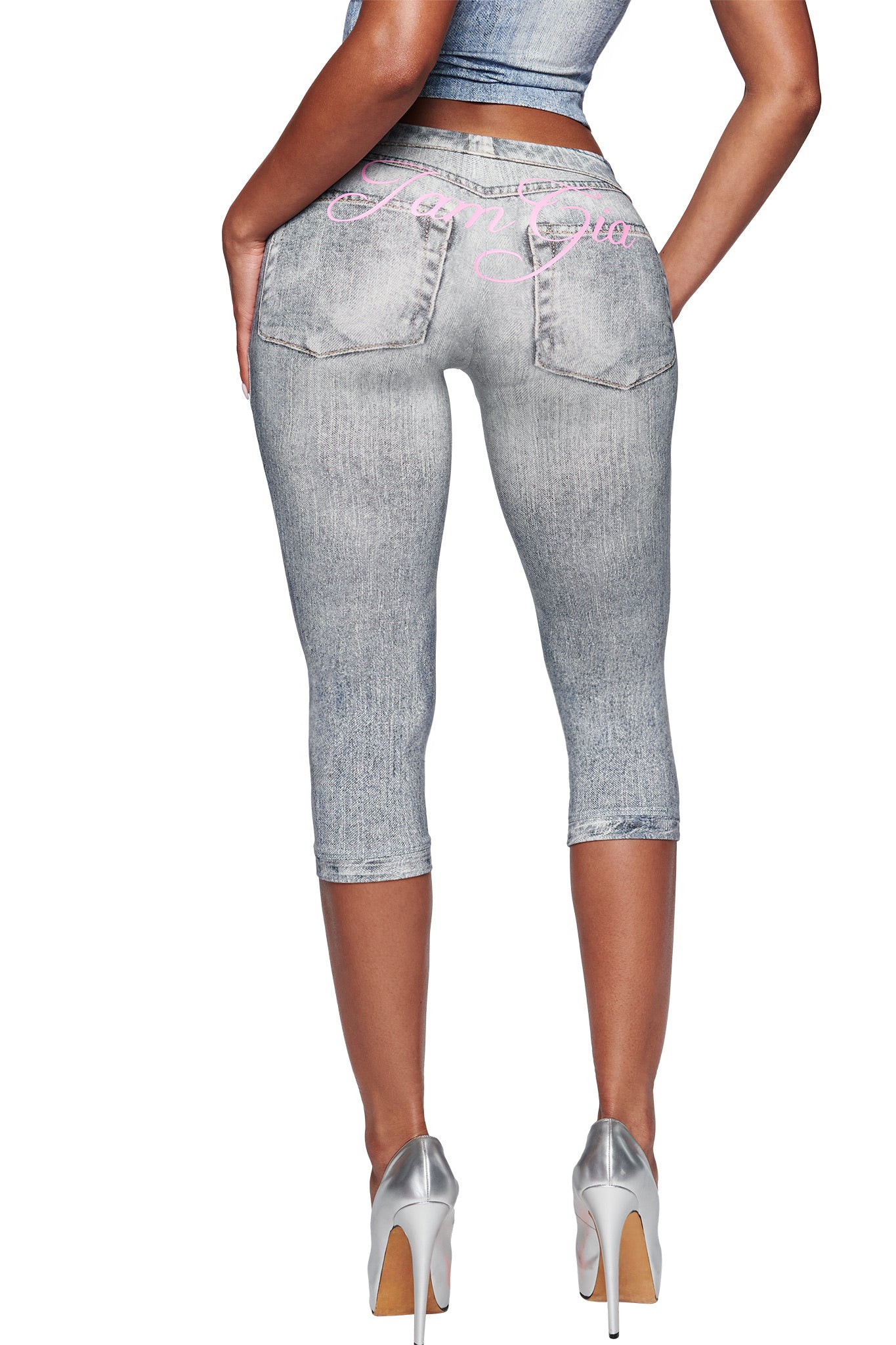 A woman is seen from the back wearing I.AM.GIA's BLARE CAPRI - DENIM TROMPE L'OEIL pants with pink cursive accents and back pockets, paired with silver heels and a matching gray top for a chic look.