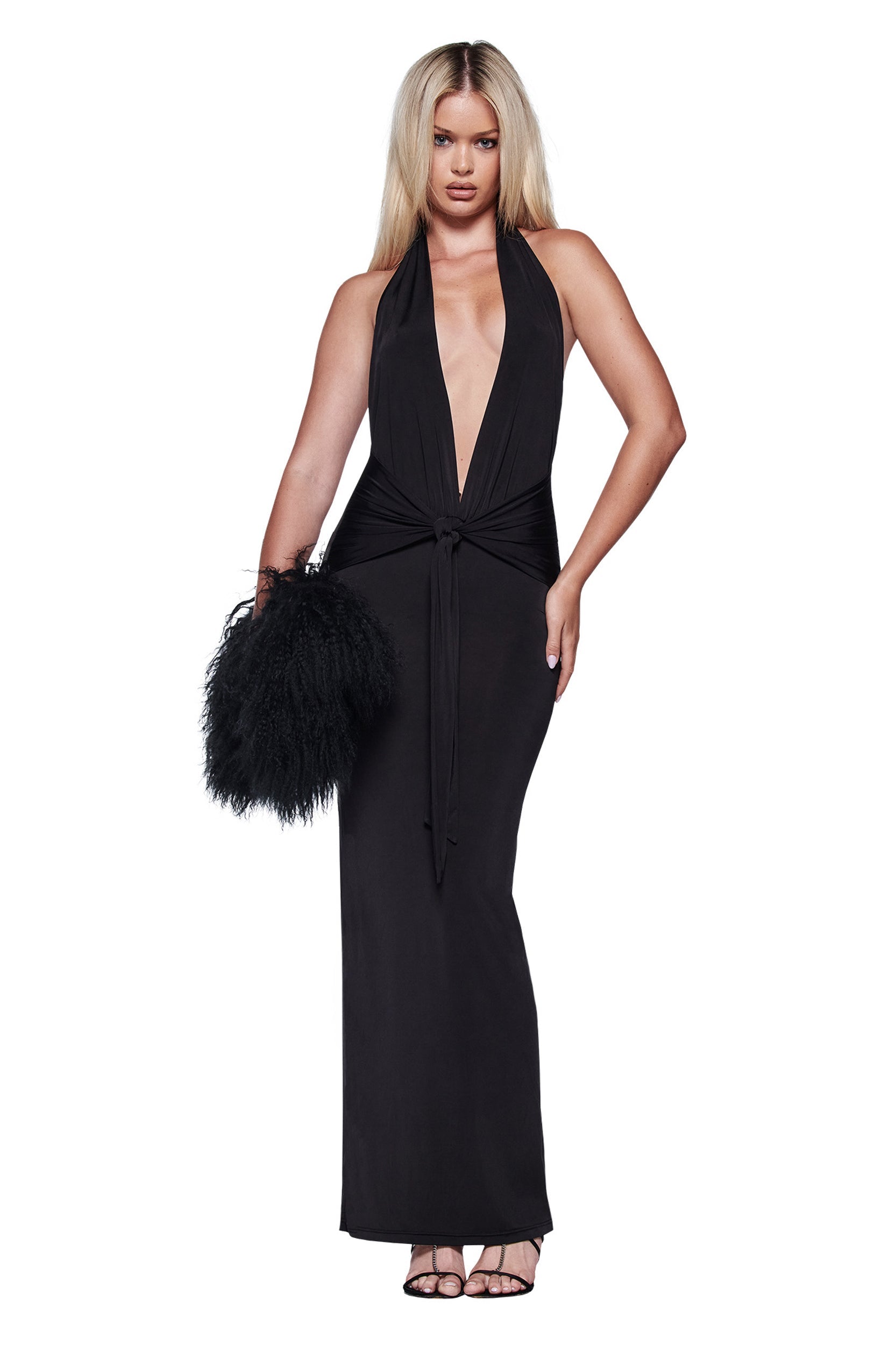 A woman wears the I.AM.GIA ZIVA PLUNGE TIE MAXI DRESS in black, featuring a deep V neckline and tie waist, paired with a feathery handbag and black strappy heels, standing against a white background.