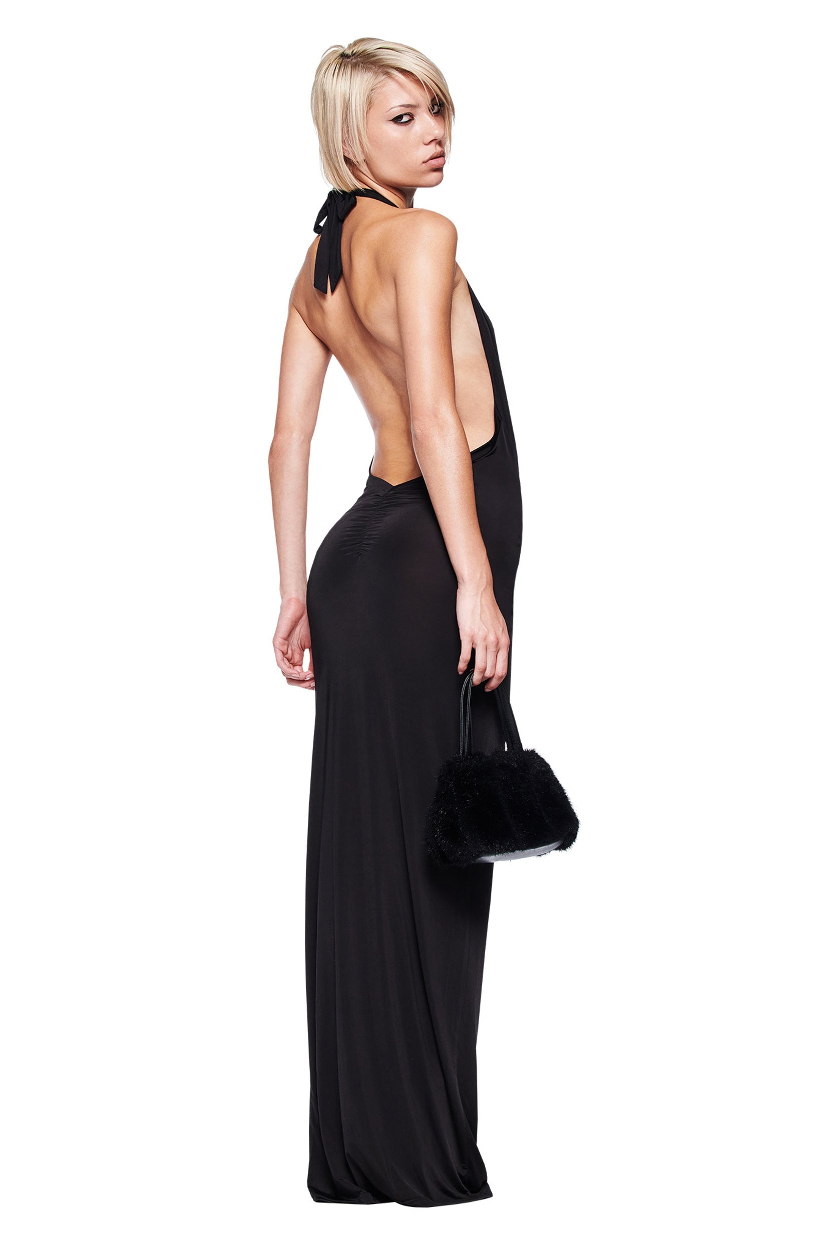 A woman with short blonde hair poses sideways, glancing over her shoulder while holding a small black bag. She wears the I.AM.GIA ZIVA PLUNGE TIE MAXI DRESS in black, featuring a plunging V neckline and bodycon fit, against a white background.