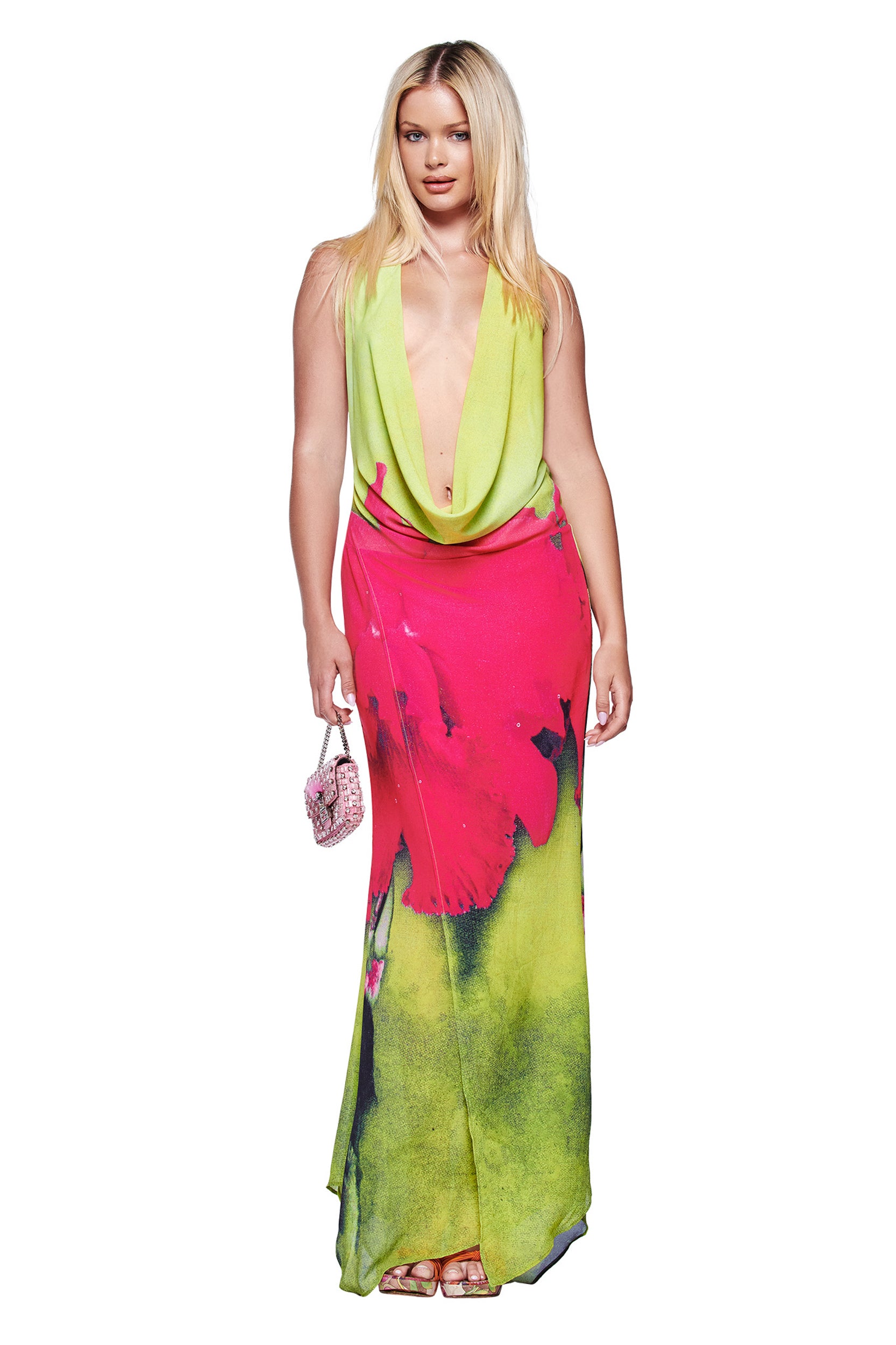 A woman wears the I.AM.GIA SAMANTHA MAXI DRESS - ORCHID PRINT, featuring a plunging cowl neck, lime green top, and pink-green skirt with neon orchid print, holding a small pink purse against a white background.