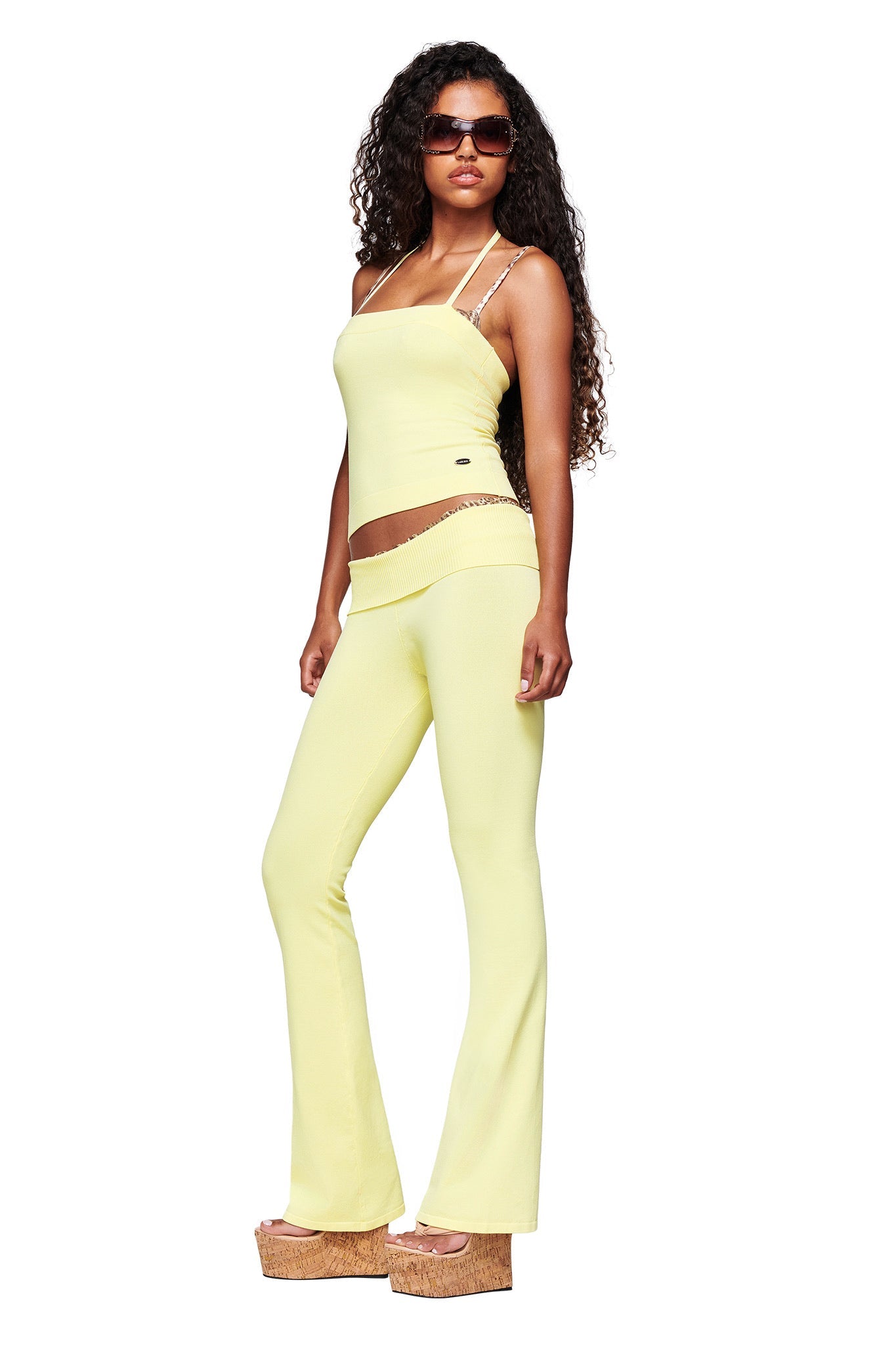 A woman in I.AM.GIA's MORGANA 2.0 PANT - BABY YELLOW and a matching tube top, styled with sunglasses and brown platform sandals, stands slightly turned to the side against a white background.
