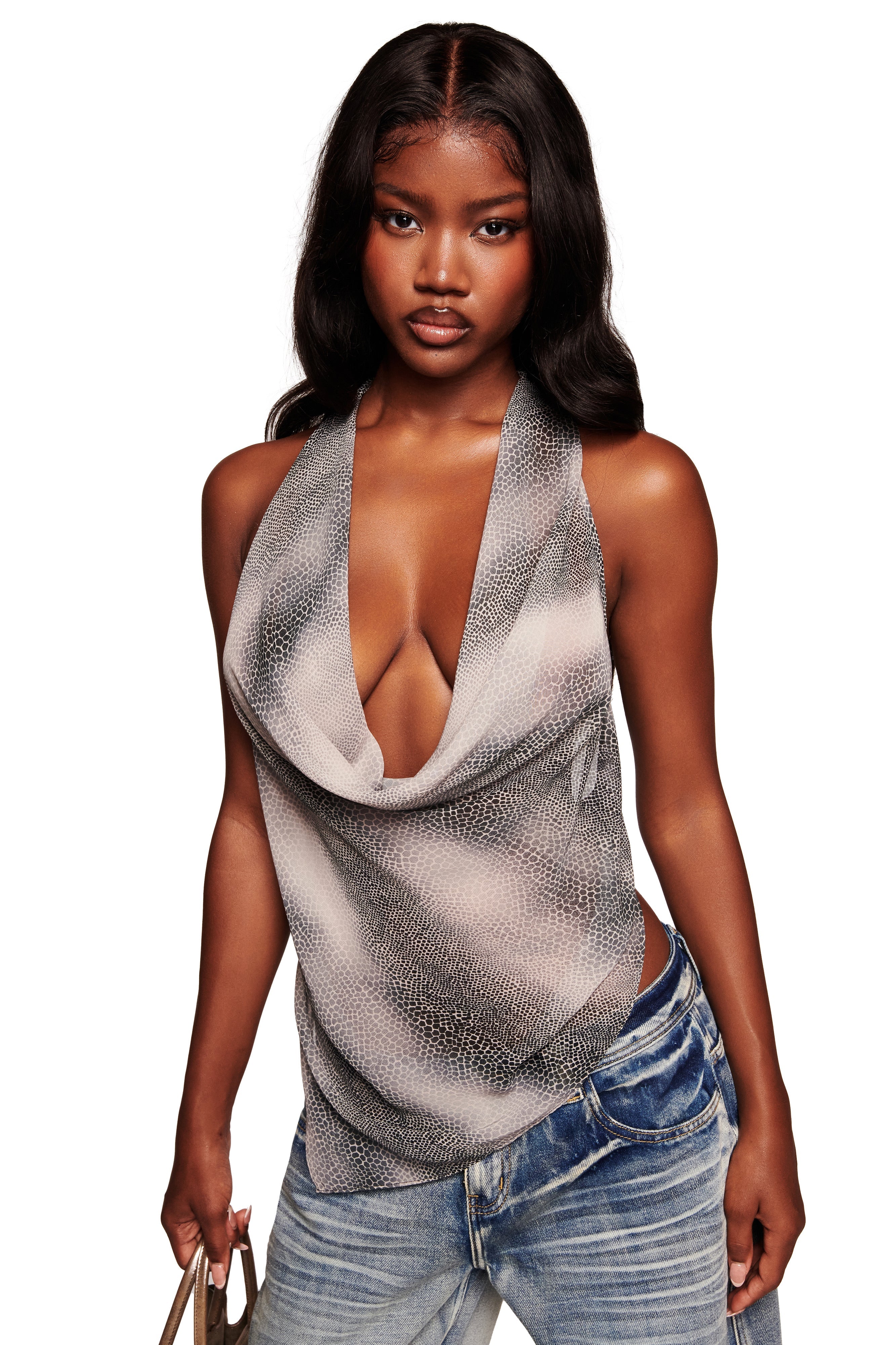 A woman with long, wavy dark hair wears the I.AM.GIA GRAYSON TOP - SERPICO, a cowl neck backless top in a black-and-white pattern, paired with low-rise blue jeans and holding a small purse. She stands against a plain white background.