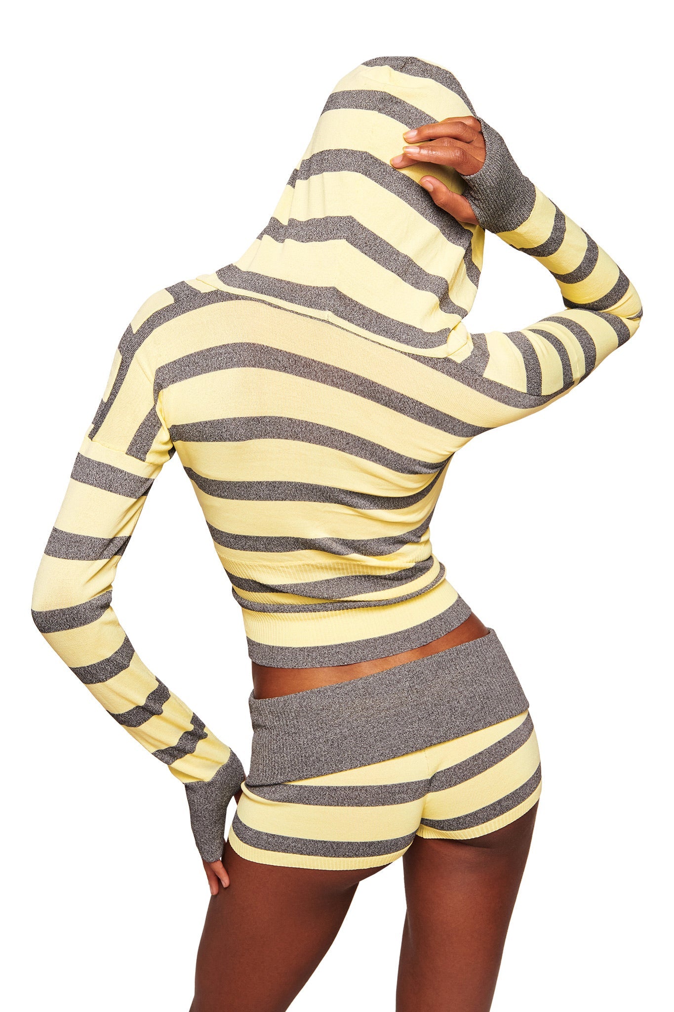 A person stands with their back to the camera, wearing the I.AM.GIA SUZI SWEATER - YELLOW AND GREY STRIPE, with matching shorts. One hand rests on their hip as they adjust the hood.