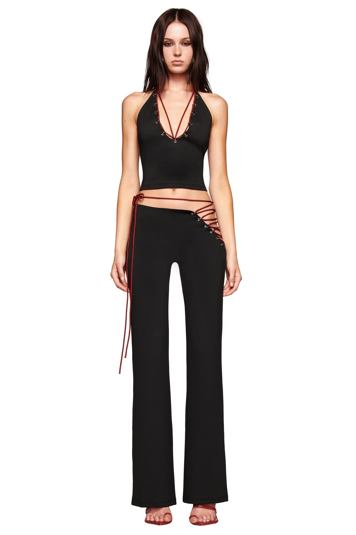 A woman faces forward barefoot against a white background, wearing I.AM.GIA's ONYX PANT - BLACK featuring cutout hip details and red lace-up sides, styled with a black halter crop top with red string accents.