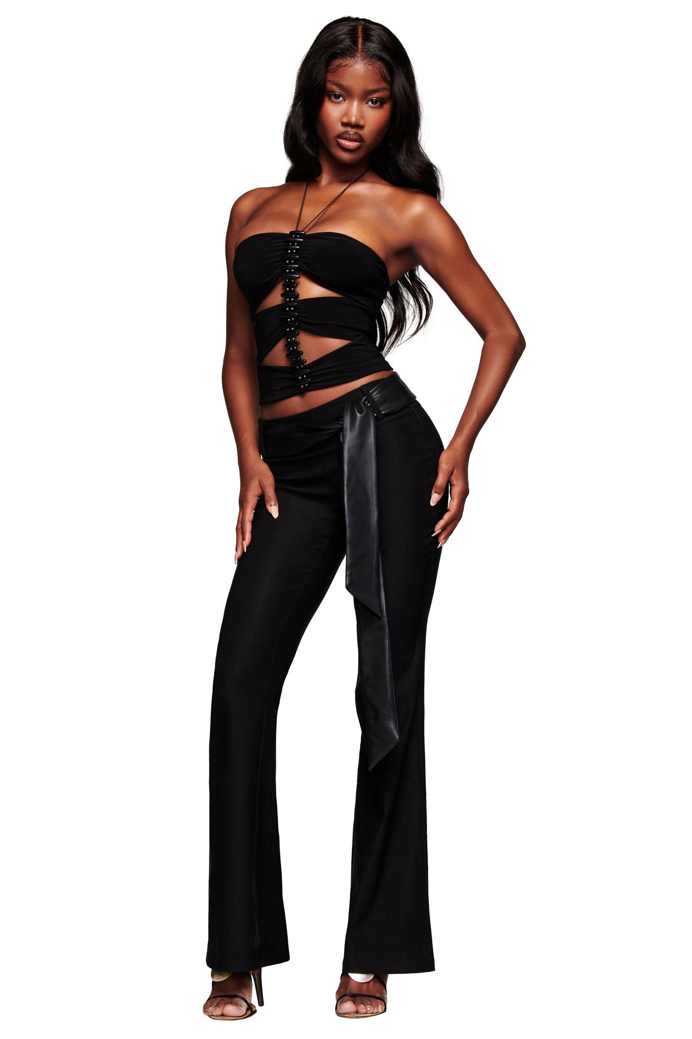 A woman poses confidently in the I.AM.GIA FENNICK TOP - BLACK with cutout details and matching high-waisted pants with a satin belt, paired with high-heeled sandals. Her long, wavy hair is loose against a white background.