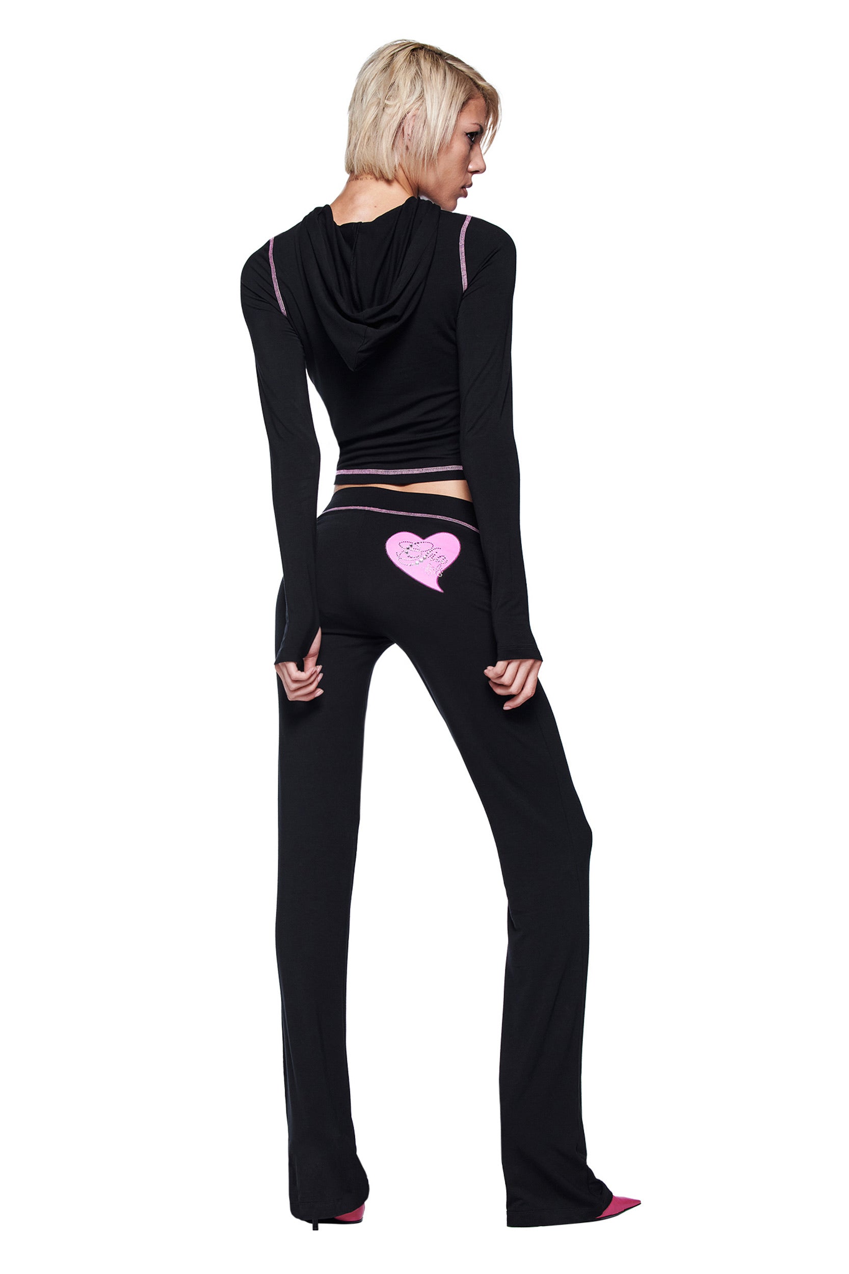 A person with short blonde hair wears a black hooded crop top and I.AM.GIA’s FAFI BLARE PANT - BLACK, which features a pink heart design on the back, standing and looking over their shoulder against a white background.