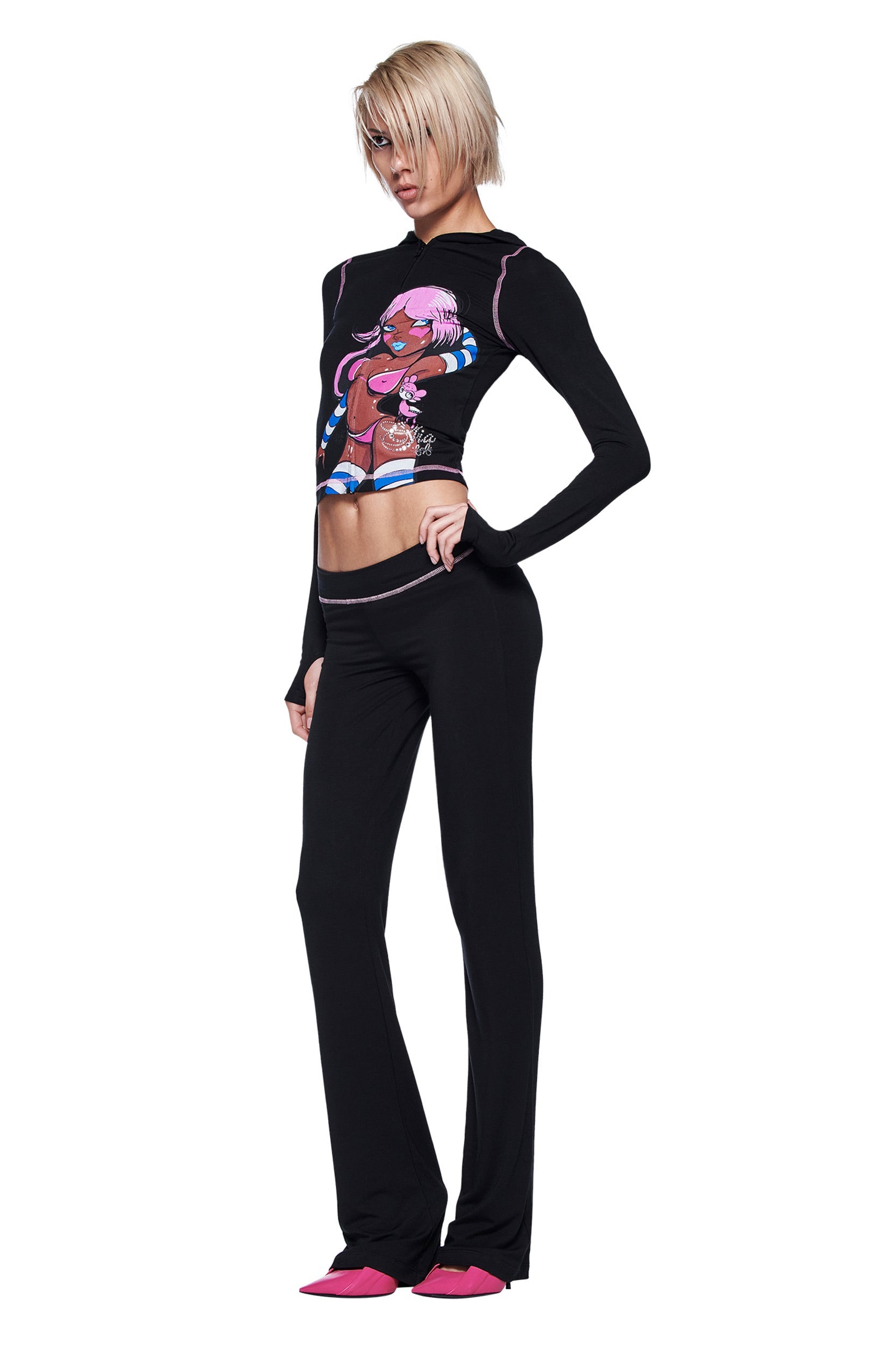 A person with short blonde hair wears the I.AM.GIA FAFI BLARE HOODIE in black, paired with black pants and pink pointed shoes. They pose against a white background with one hand on their hip.