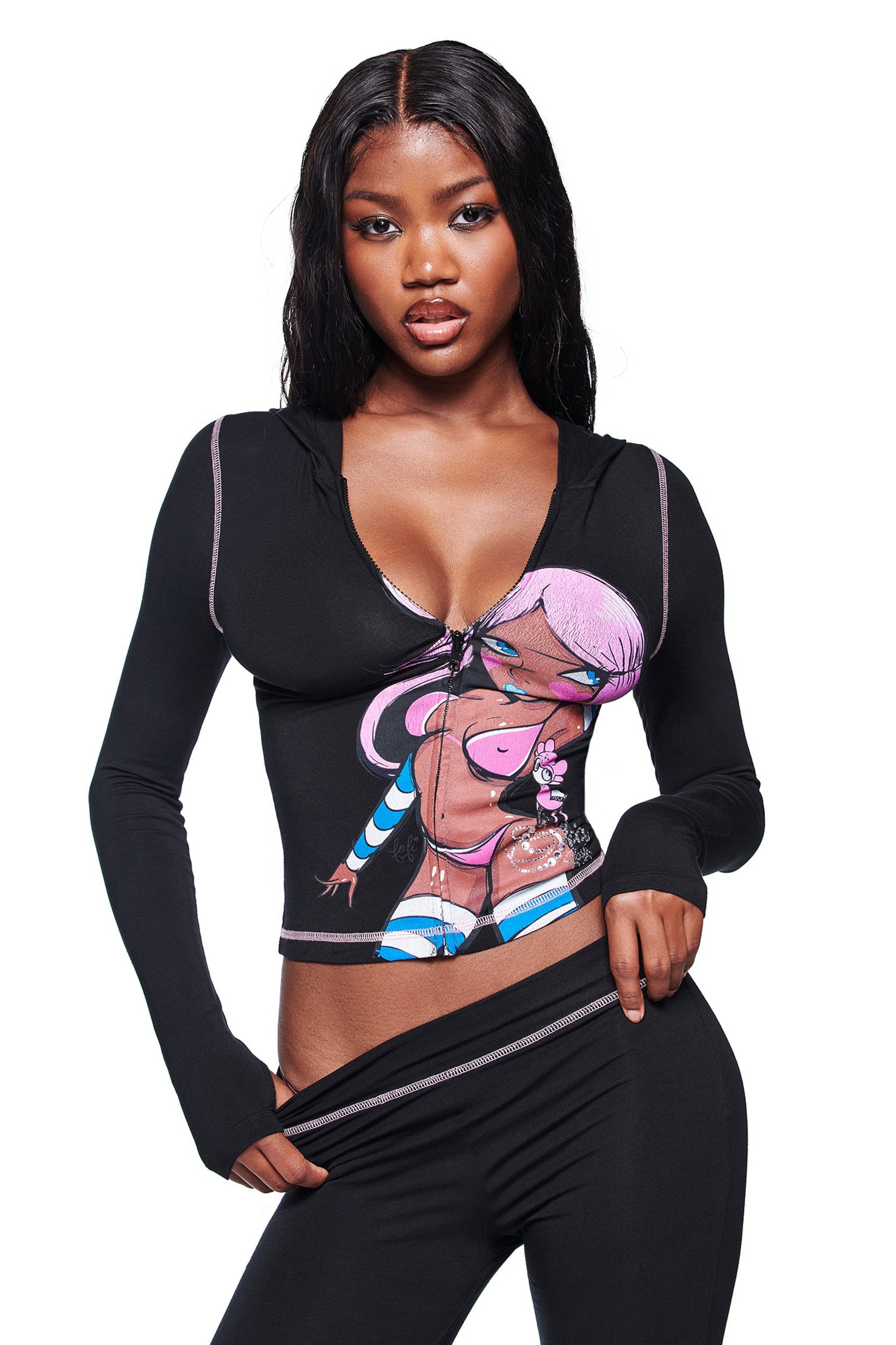 A woman with long, wavy black hair wears the I.AM.GIA FAFI BLARE HOODIE in black, featuring a pink-haired cartoon graphic from the Fafi collaboration. She poses against a white background, pulling at her waistband.