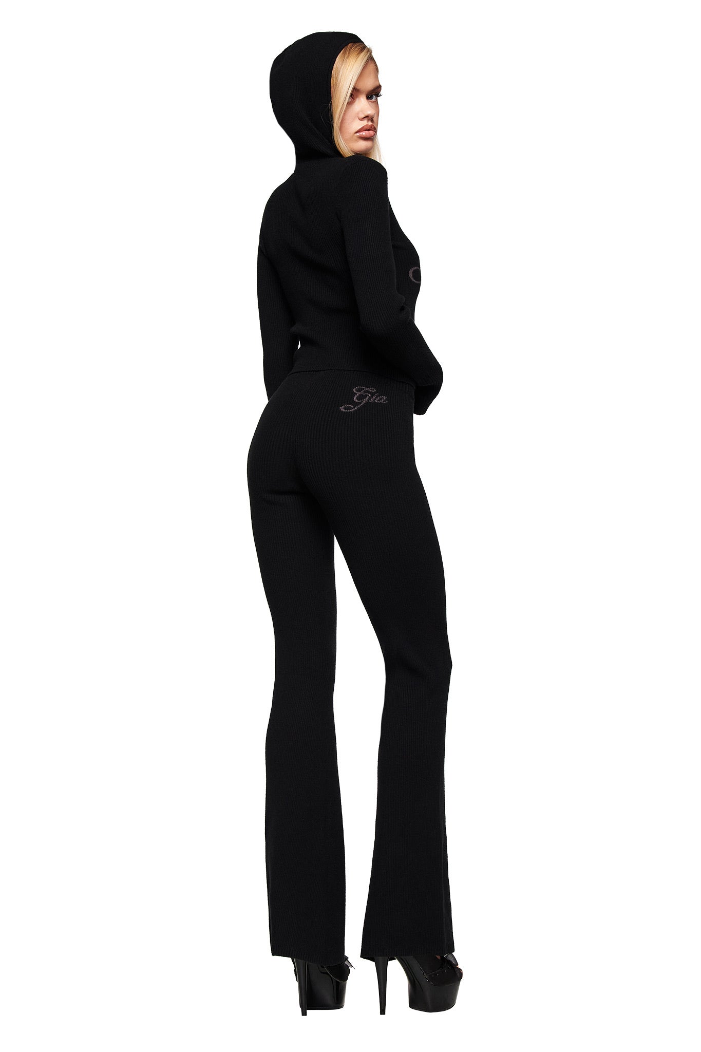 A woman in the I.AM.GIA BLARE KNIT TRACKPANT - BLACK stands turned to the side, wearing a fitted black hooded sweater and black platform heels, looking over her shoulder against a white background.