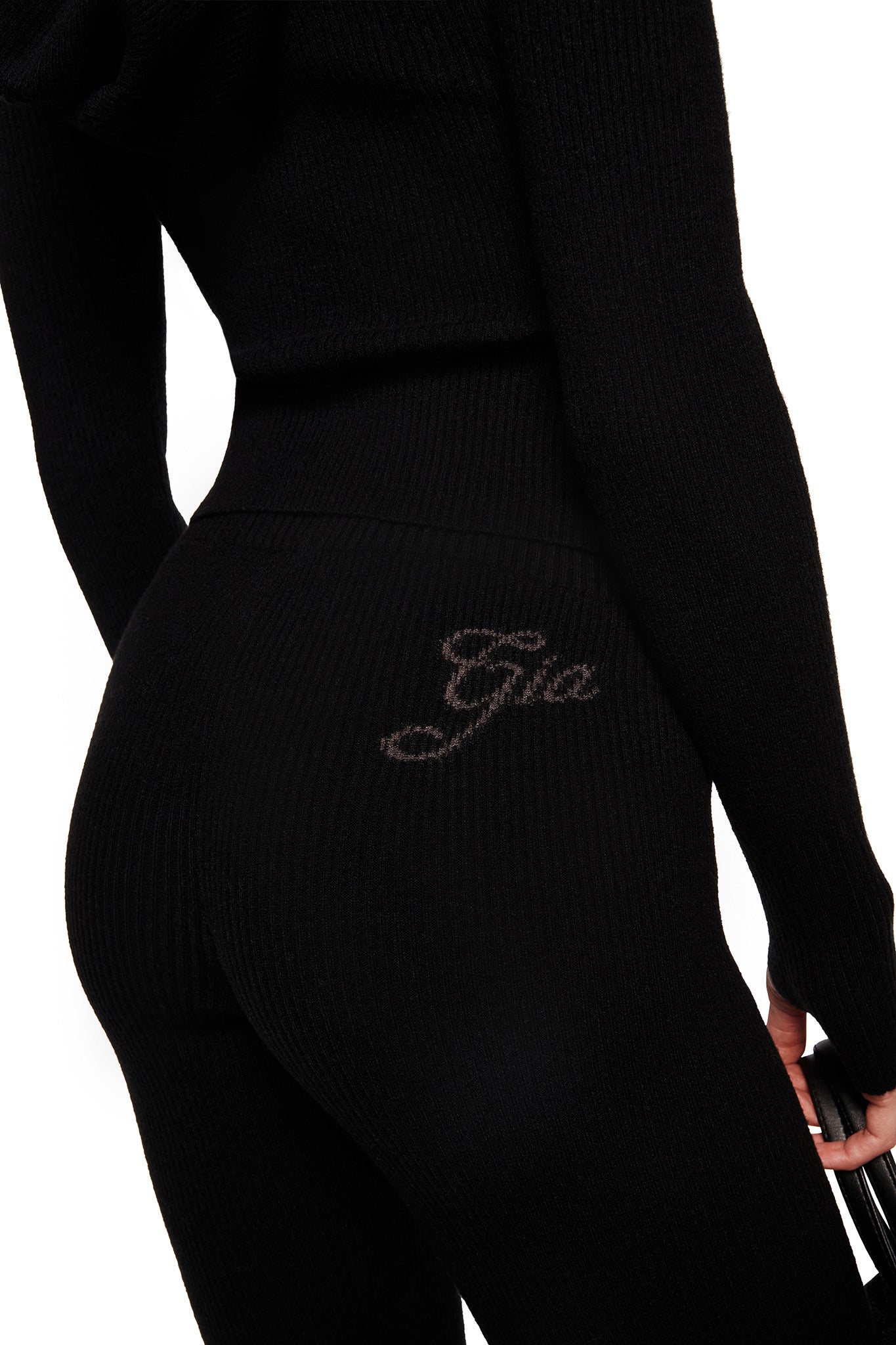 A person wears a fitted black ribbed long-sleeve top and I.AM.GIA BLARE KNIT TRACKPANT in black, featuring a mid-rise elastic waistband, "Gia" embroidery on the hip, and flared leg opening, holding a small item in hand.