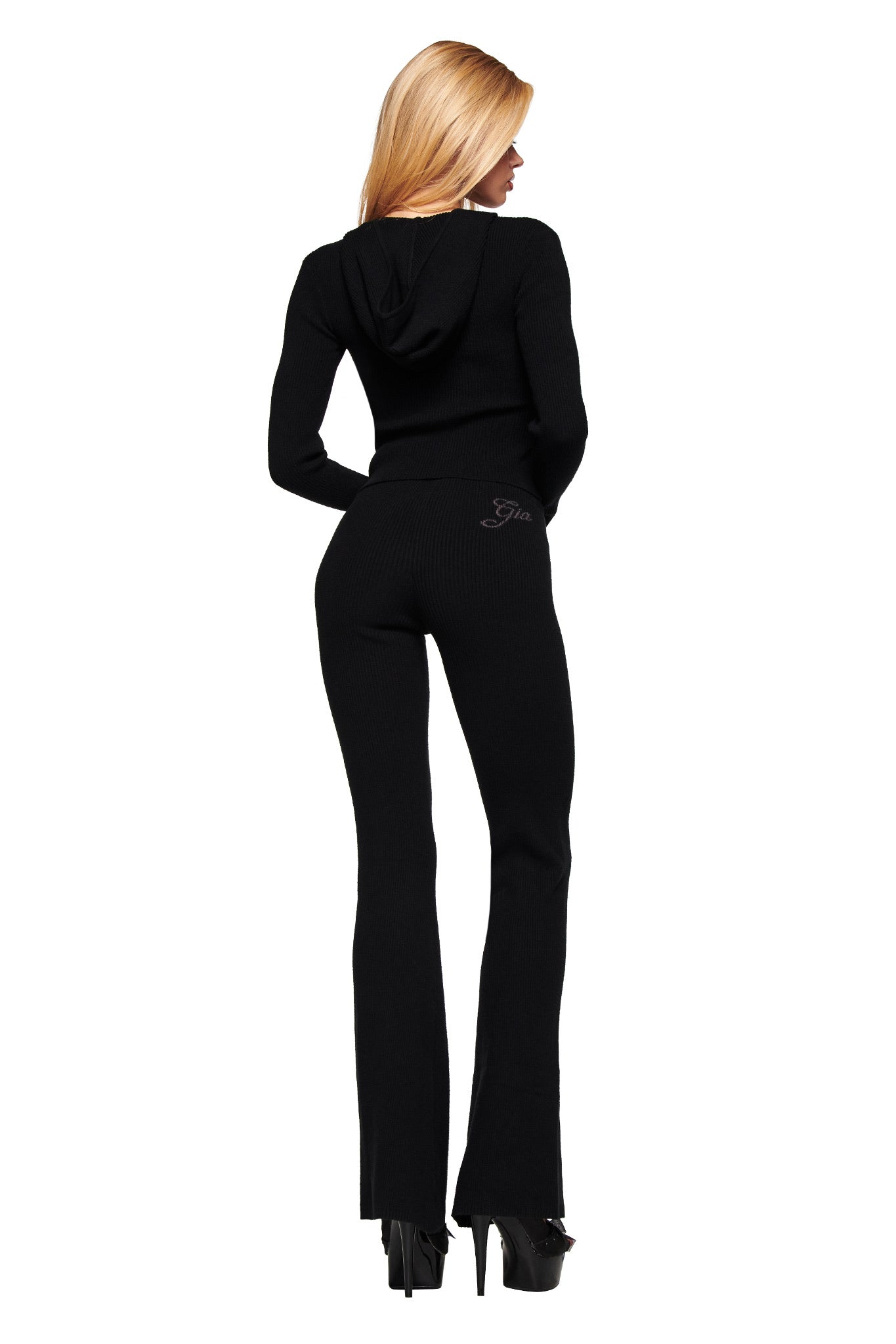 A woman with long blonde hair faces away, wearing the I.AM.GIA BLARE KNIT HOODIE in black with a bodycon fit and matching flared pants plus platform heels, set against a white background.