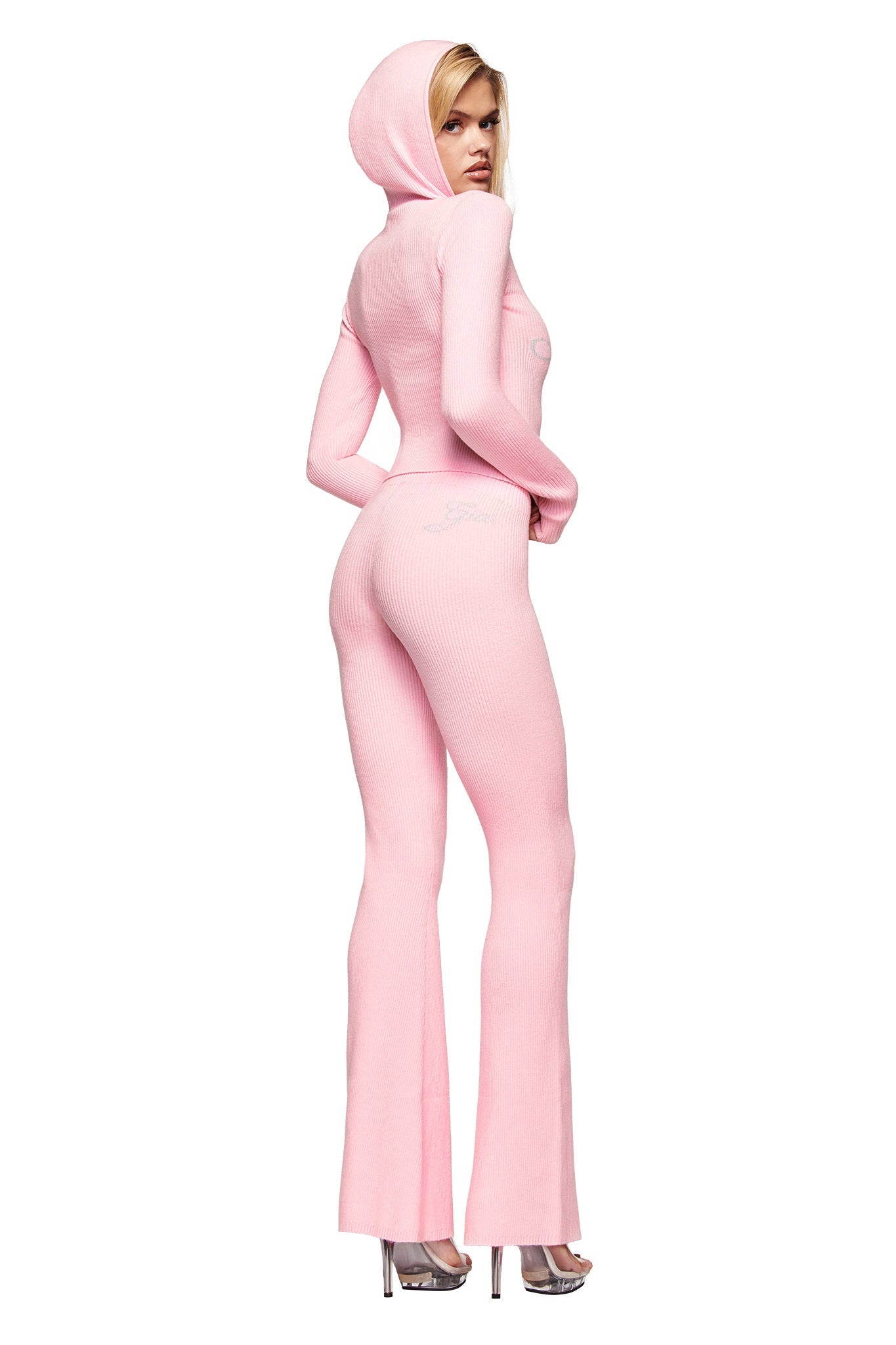 A woman models the I.AM.GIA BLARE KNIT HOODIE in pink—a fitted knit jumpsuit with flared legs and a ribbed texture—paired with clear heels, posing against a white background.
