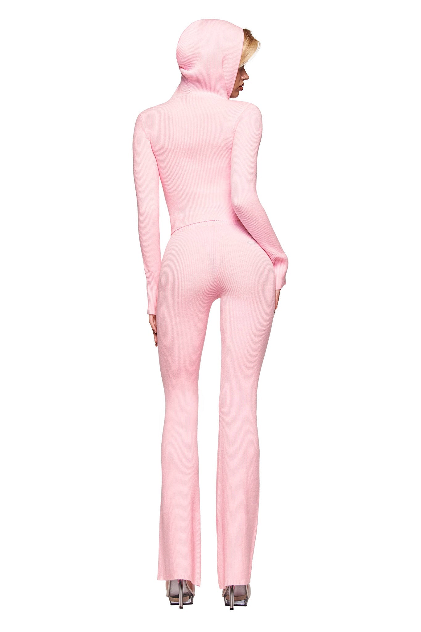A person faces away, wearing I.AM.GIA’s BLARE KNIT TRACKPANT in pink styled with a fitted light pink hooded jumpsuit and clear high heels against a white background.