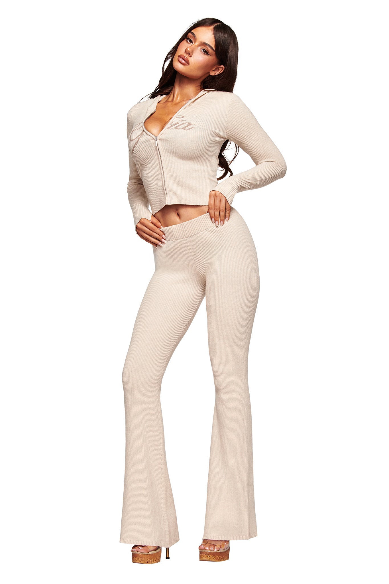 A woman models the I.AM.GIA BLARE KNIT TRACKPANT in cream, featuring a mid rise elasticated waistband and flared leg, styled with a matching beige jacket and nude platform heels against a white background.