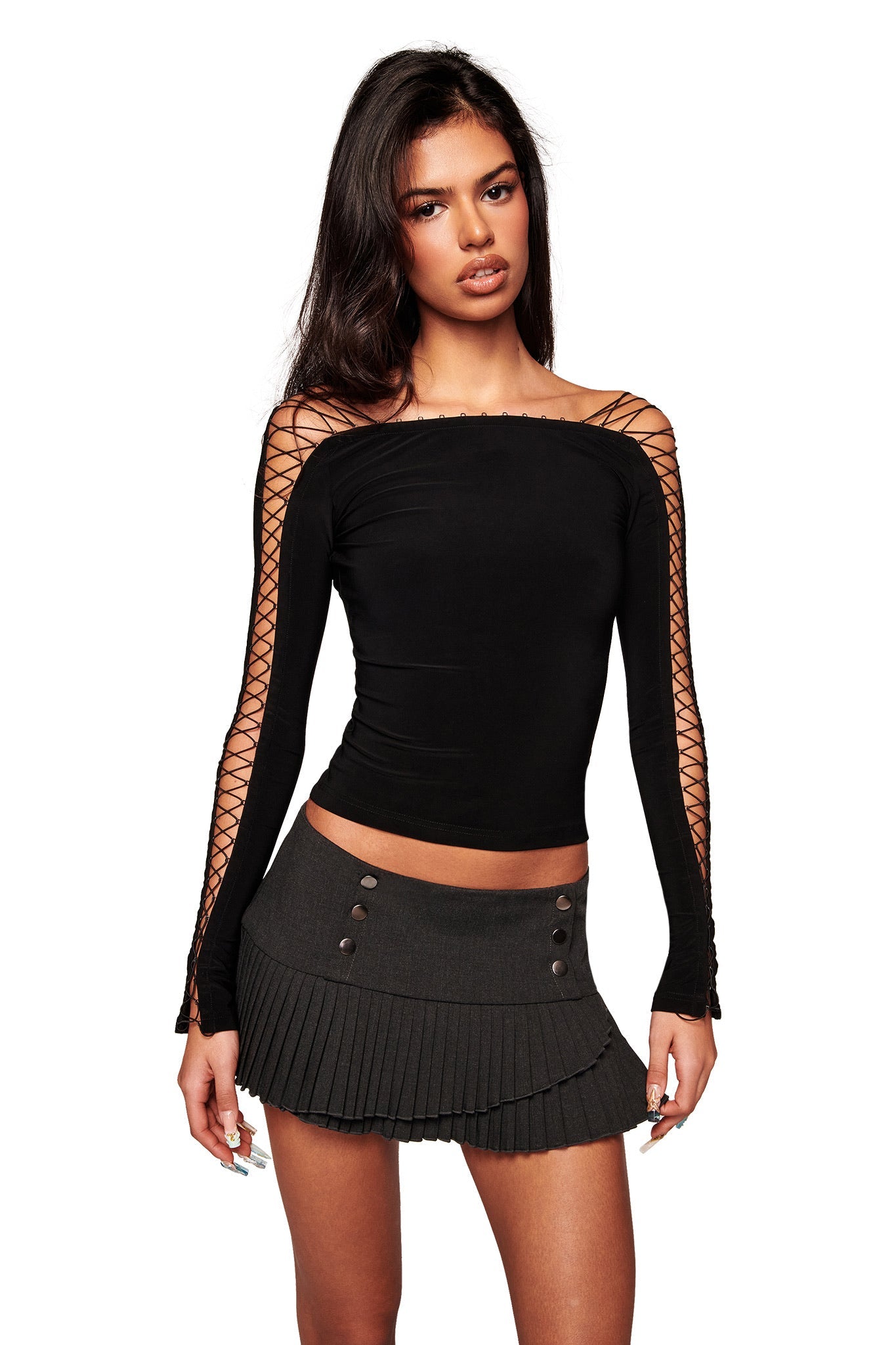 A woman wears the I.AM.GIA VERO TOP in black, a lace-up long sleeve bodycon piece with crisscross details, paired with a dark gray pleated mini skirt featuring button accents, standing against a white background.