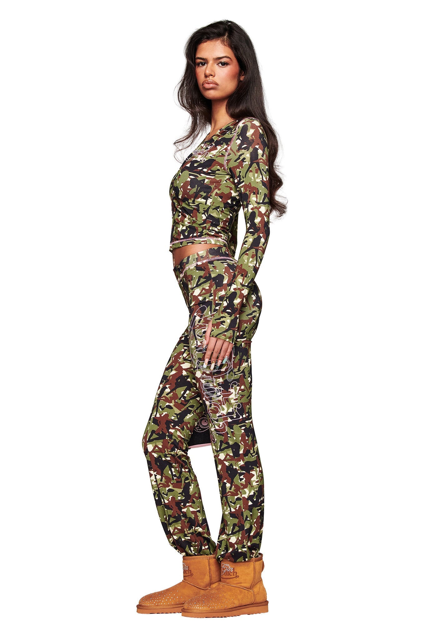 Woman in a side pose wears an I.AM.GIA X VON DUTCH BLARE TRACKPANT – STRIPPER CAMO, a long-sleeve camo top, and tan Timberland-style boots against a white background.