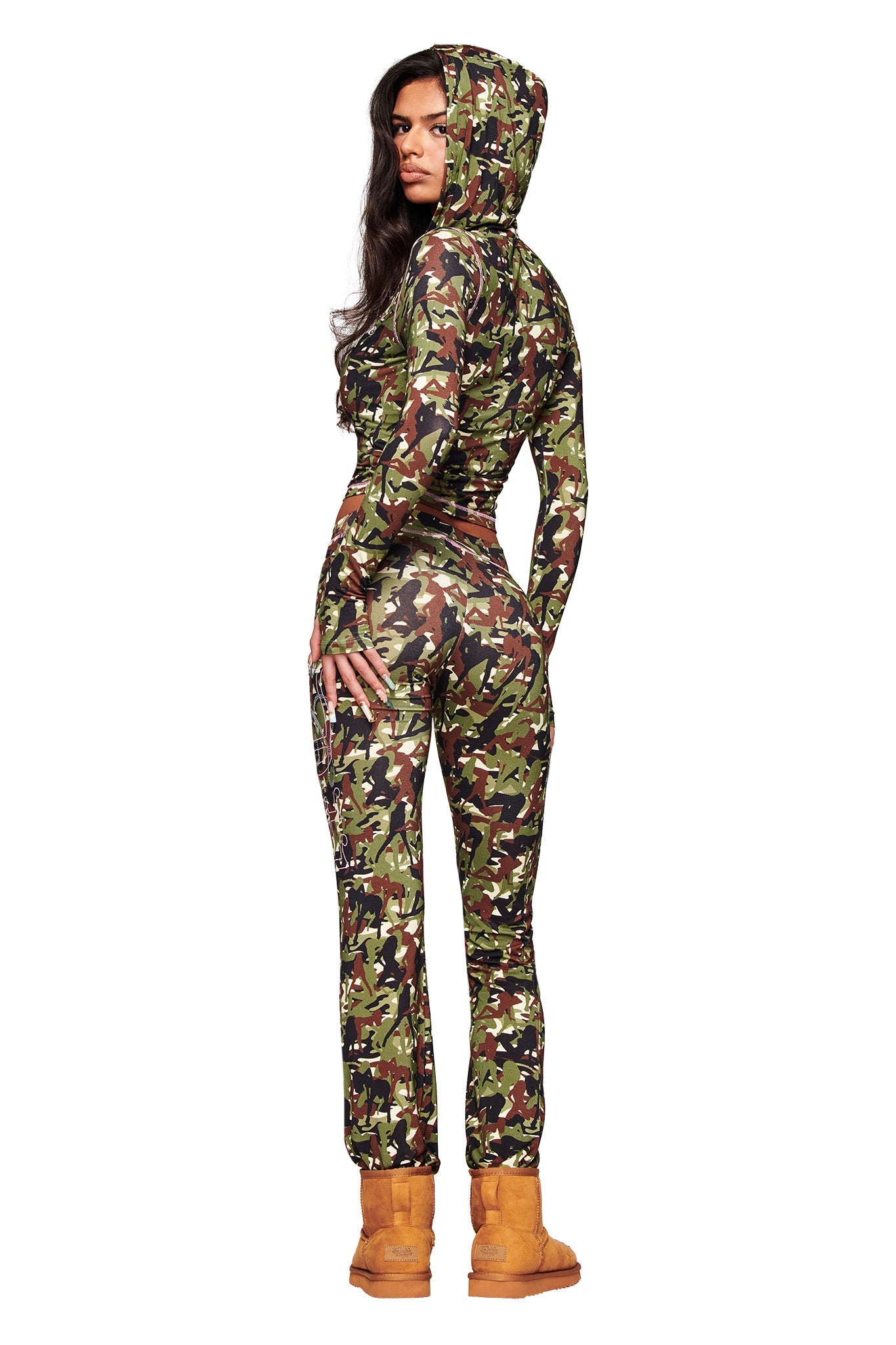 A woman with long, dark hair poses confidently against a white background in the I.AM.GIA X VON DUTCH Blare Trackpant - Stripper Camo by I.AM.GIA, styled with tan boots and featuring a bold camouflage print.