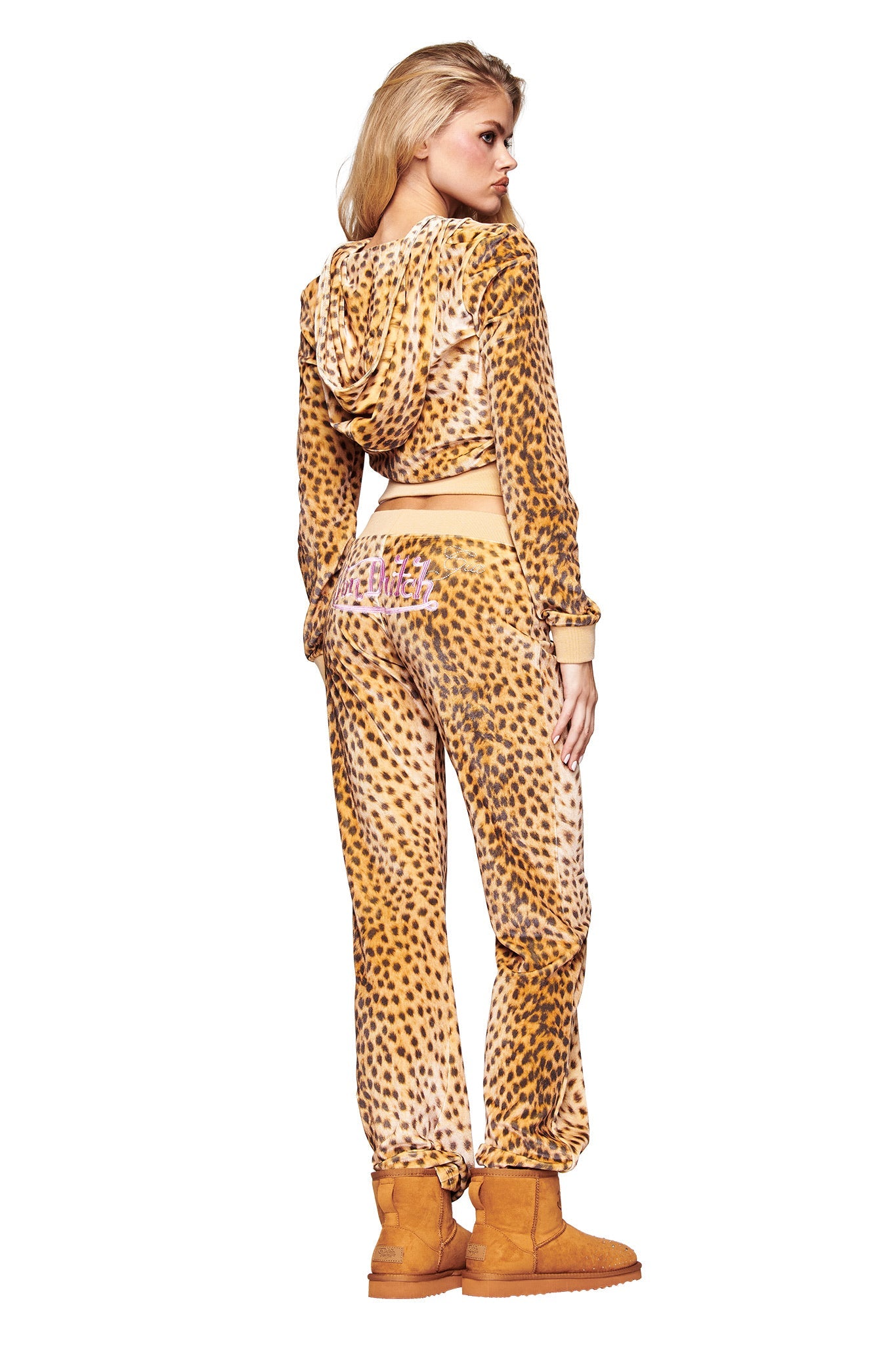A woman with long blonde hair poses confidently in the I.AM.GIA X VON DUTCH KAMERON VELOUR JACKET - LEOPARD, paired with matching pants and tan boots, facing away against a white background.