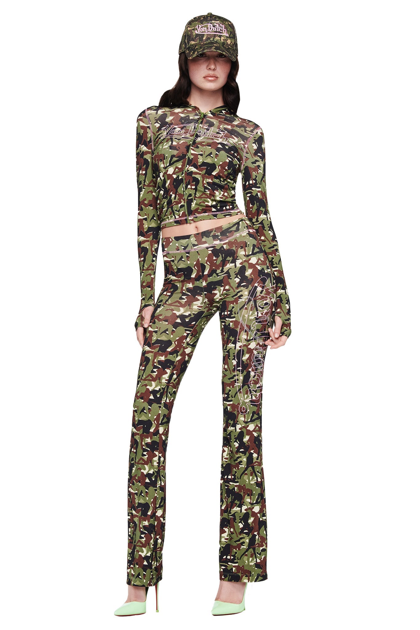 A woman stands facing forward in a matching camouflage outfit: the I.AM.GIA X Von Dutch Blare Trackpant – Gia Girl Camo, a camo crop top, and a Von Dutch cap, paired with light green high heels.