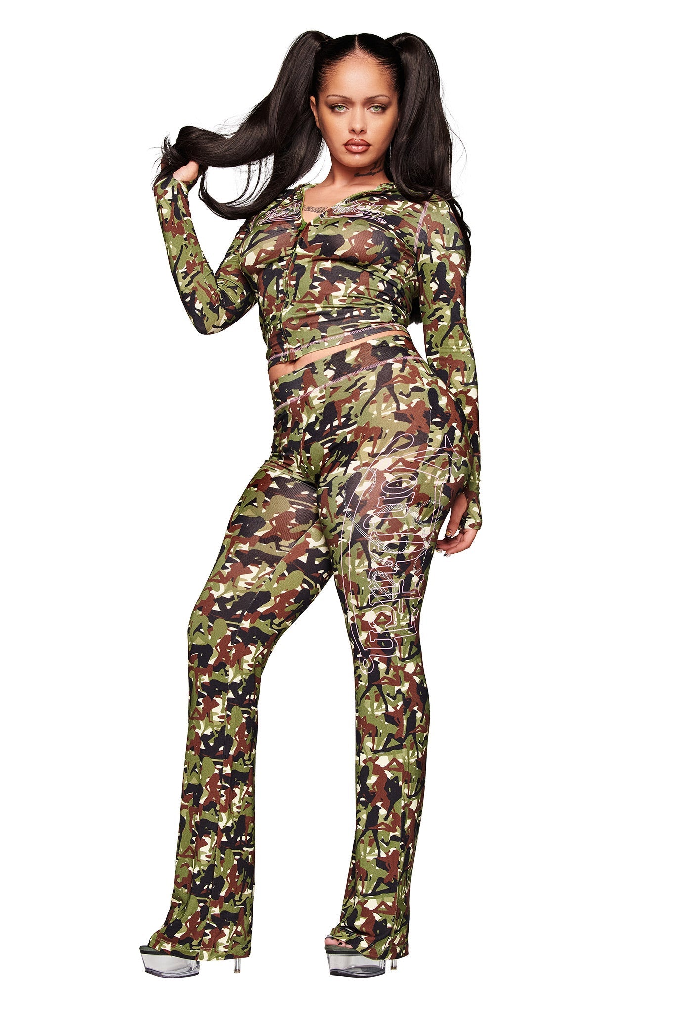 A woman with long pigtails stands confidently against a white background, wearing I.AM.GIA X VON DUTCH Blare Trackpants in Stripper Camo and a matching crop top, paired with silver high heels.
