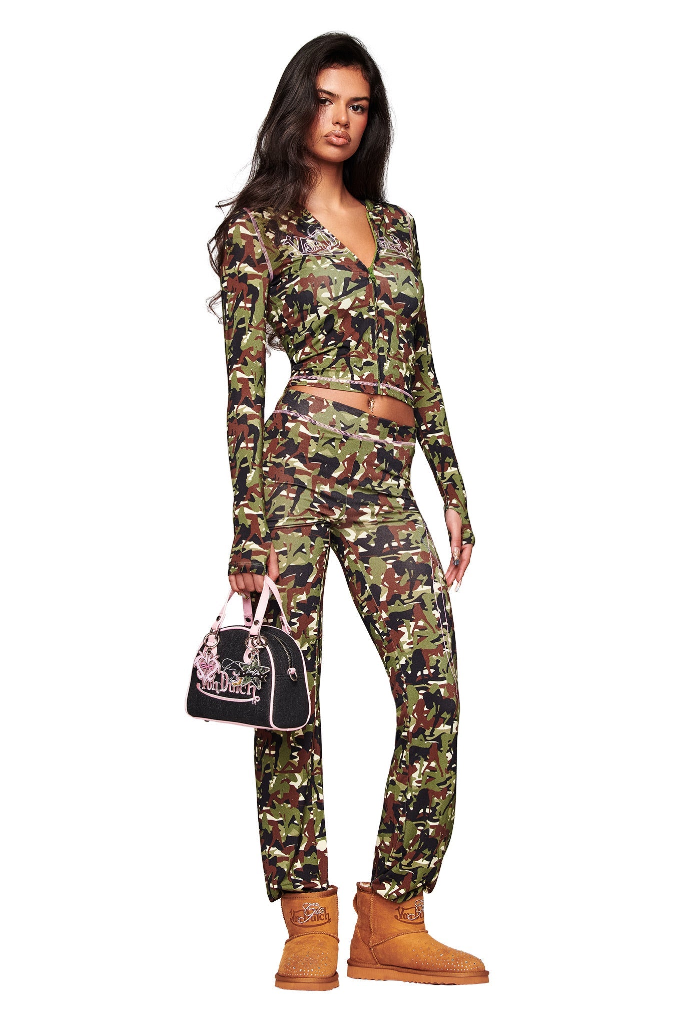 A woman faces forward in the I.AM.GIA x Von Dutch Blare Hoodie in Gia Girl Camo, styled with matching pants. She holds a black handbag, wears tan boots, and lets her long dark hair down while posing against a white background.