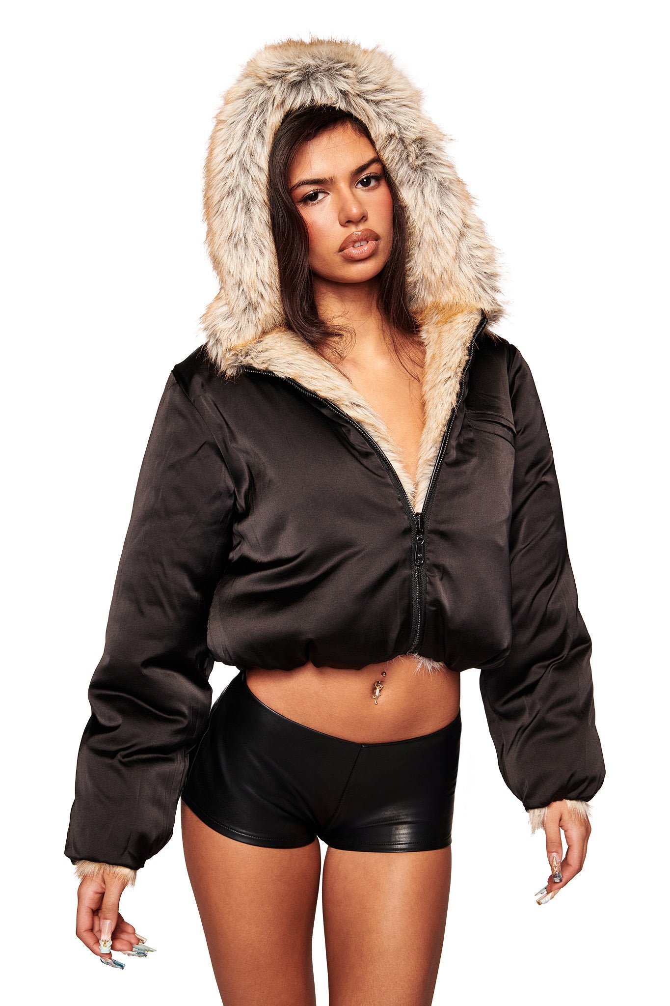 A woman wears the I.AM.GIA EVEREST REVERSIBLE JACKET in brown, featuring a faux fur-lined hood, paired with black high-waisted shorts, standing against a white background.