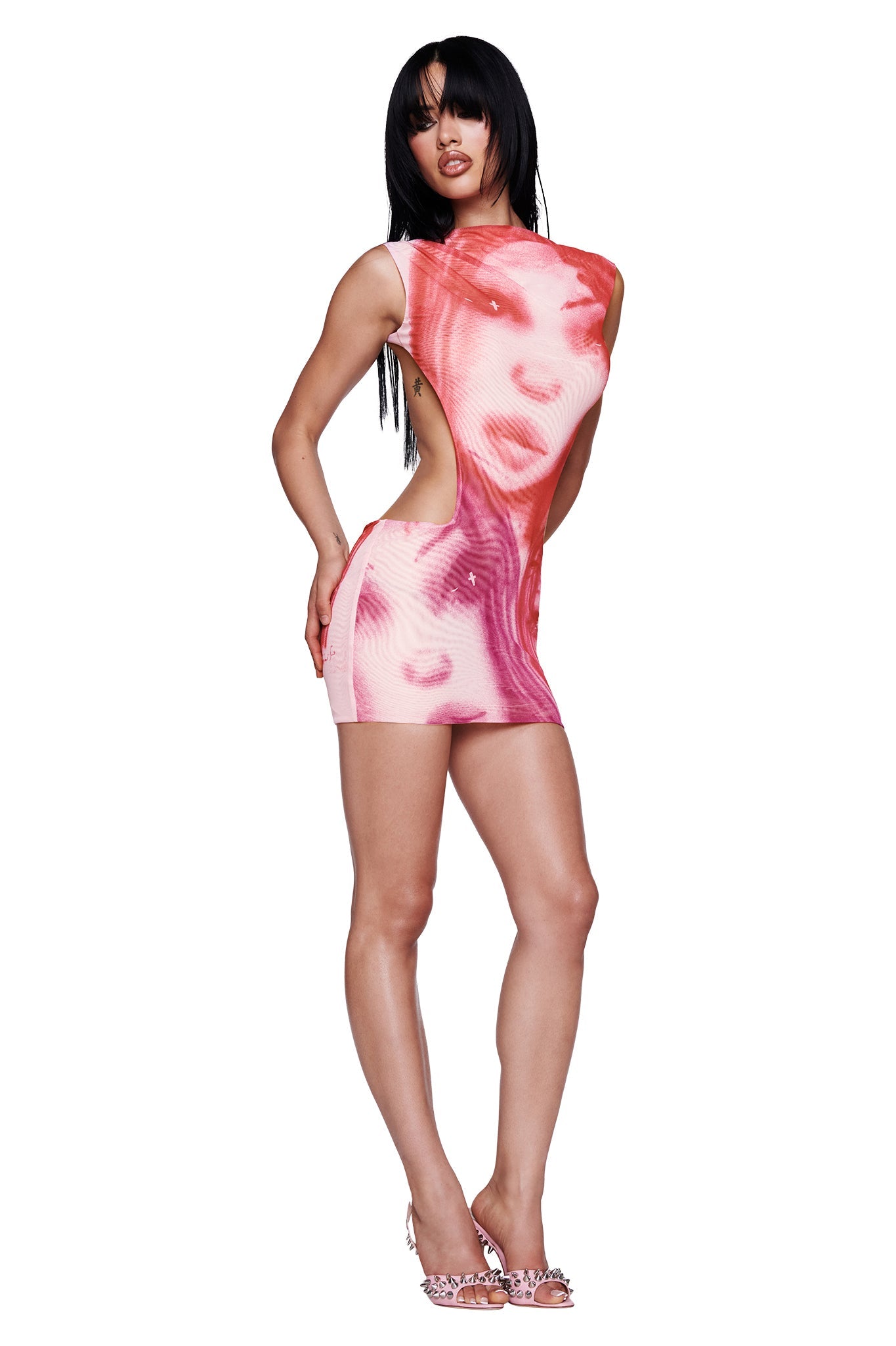 A woman with long black hair poses confidently against a white background wearing the I.AM.GIA T-REX CORALINE MINI DRESS in pink, a micro mini dress featuring abstract white face patterns and open sides, styled with high heels—the perfect festival dress.