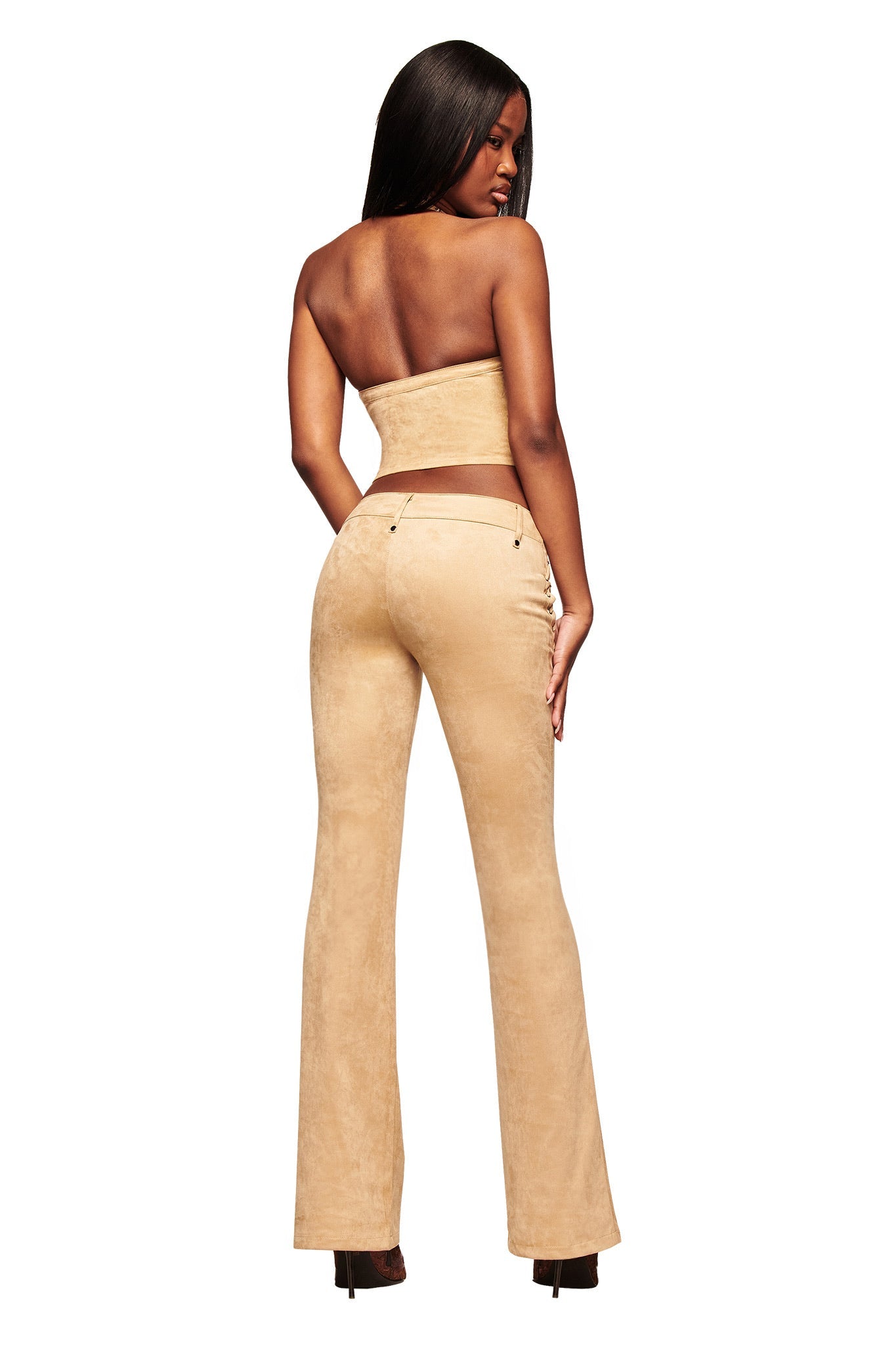 A woman with long hair poses with her back turned, wearing the I.AM.GIA XENIA SUEDE PANT in tan and a strapless beige top. She completes the look with high heels against a white background.