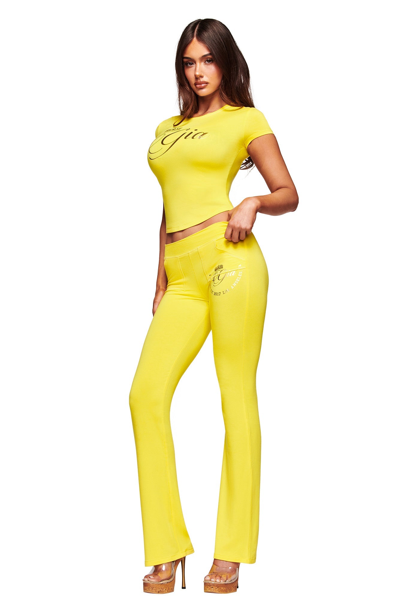 A woman wears I.AM.GIA’s BLARE FORM POCKET TRACKPANT in yellow gold foil—featuring sculpted Form fabric with cursive writing—styled with a matching crop top, tan platform heels, and long, loose hair.