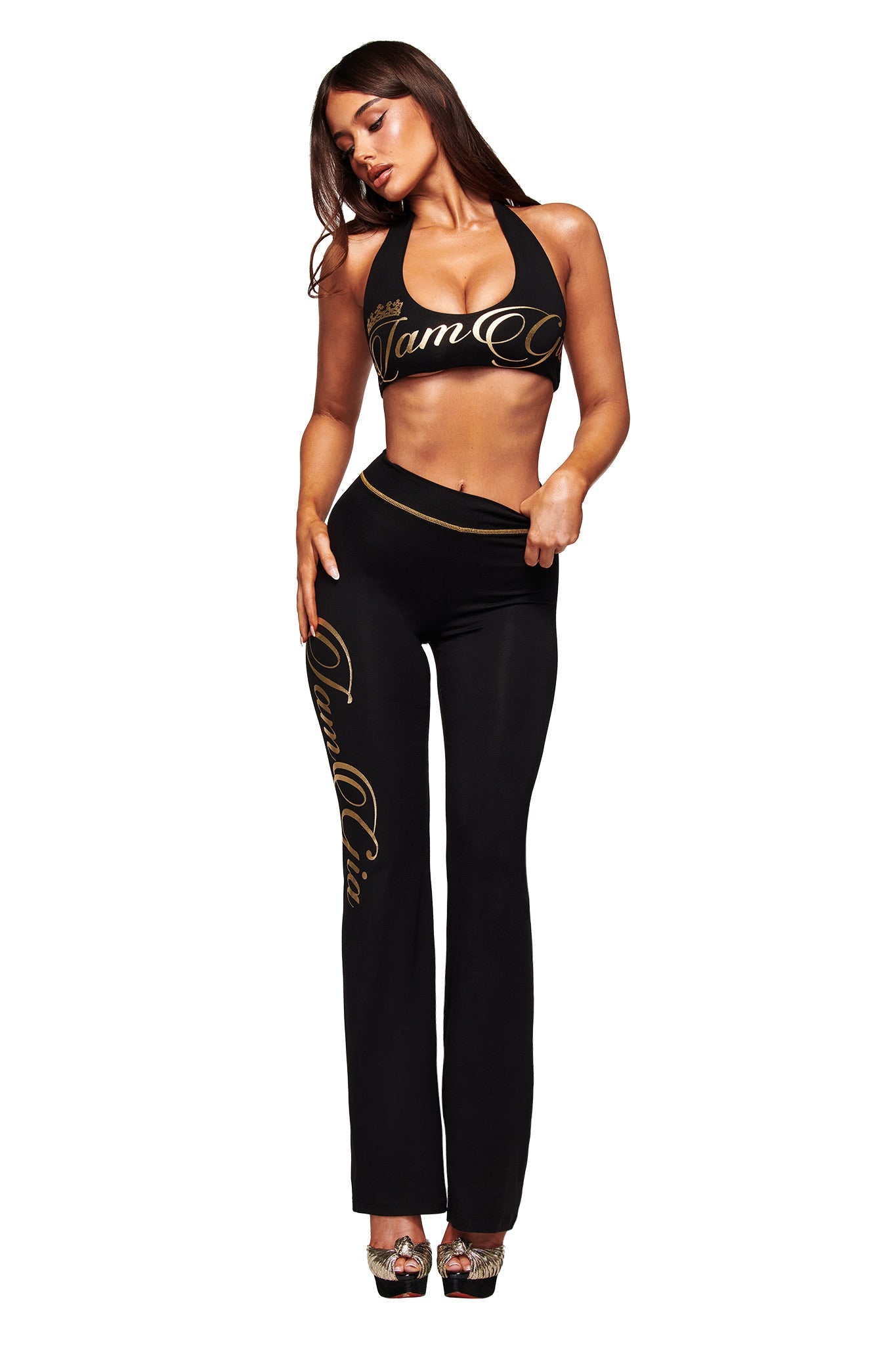 A woman poses in the I.AM.GIA BLARE FORM TRACKPANT - BLACK AND GOLD, paired with a black halter crop top and high heels. The sculpted Form fabric accentuates her silhouette against a plain white background.