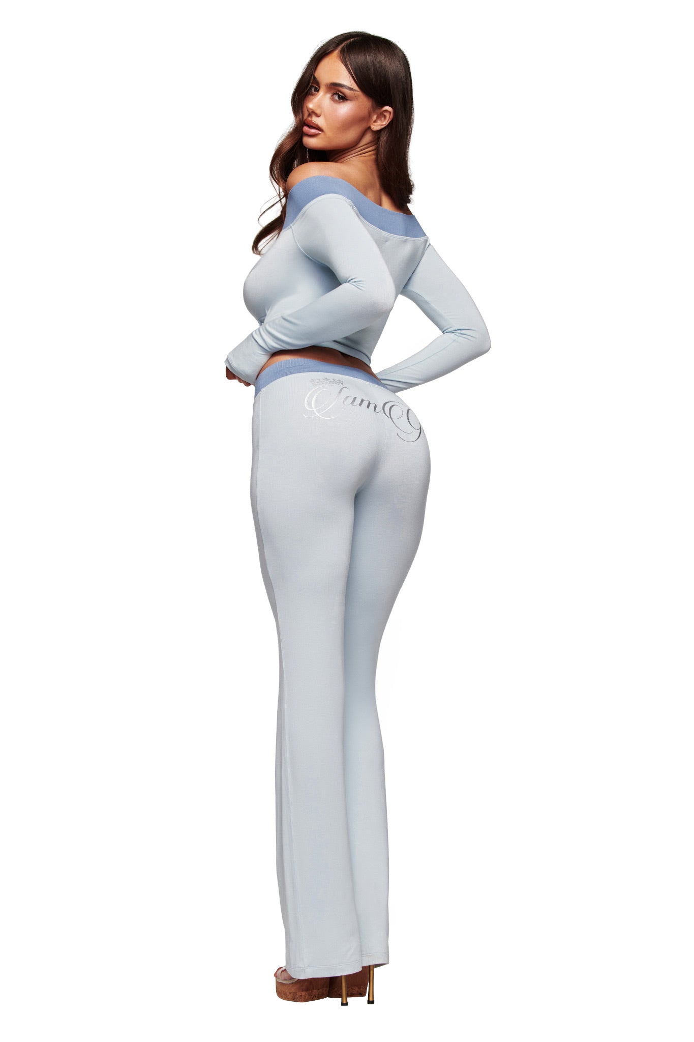 A woman with long brown hair wears the I.AM.GIA BLARE OFF SHOULDER TOP in two tone powder blue, paired with high-waisted pants. She stands with her back to the camera, glancing over her shoulder in high heels.