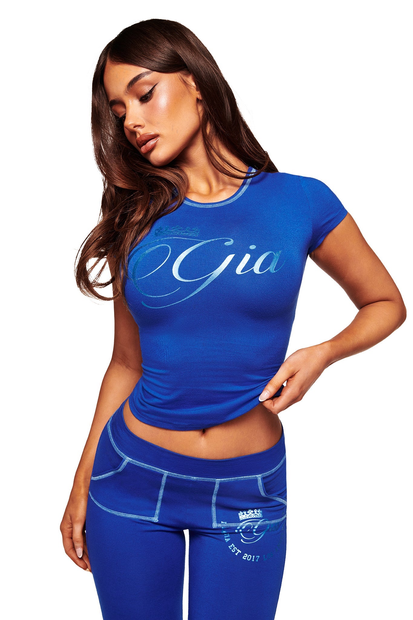 Woman with long brown hair wears the I.AM.GIA BLARE FORM TEE - COBALT FOIL and matching blue pants, posing with one hand on her hip against a white background.