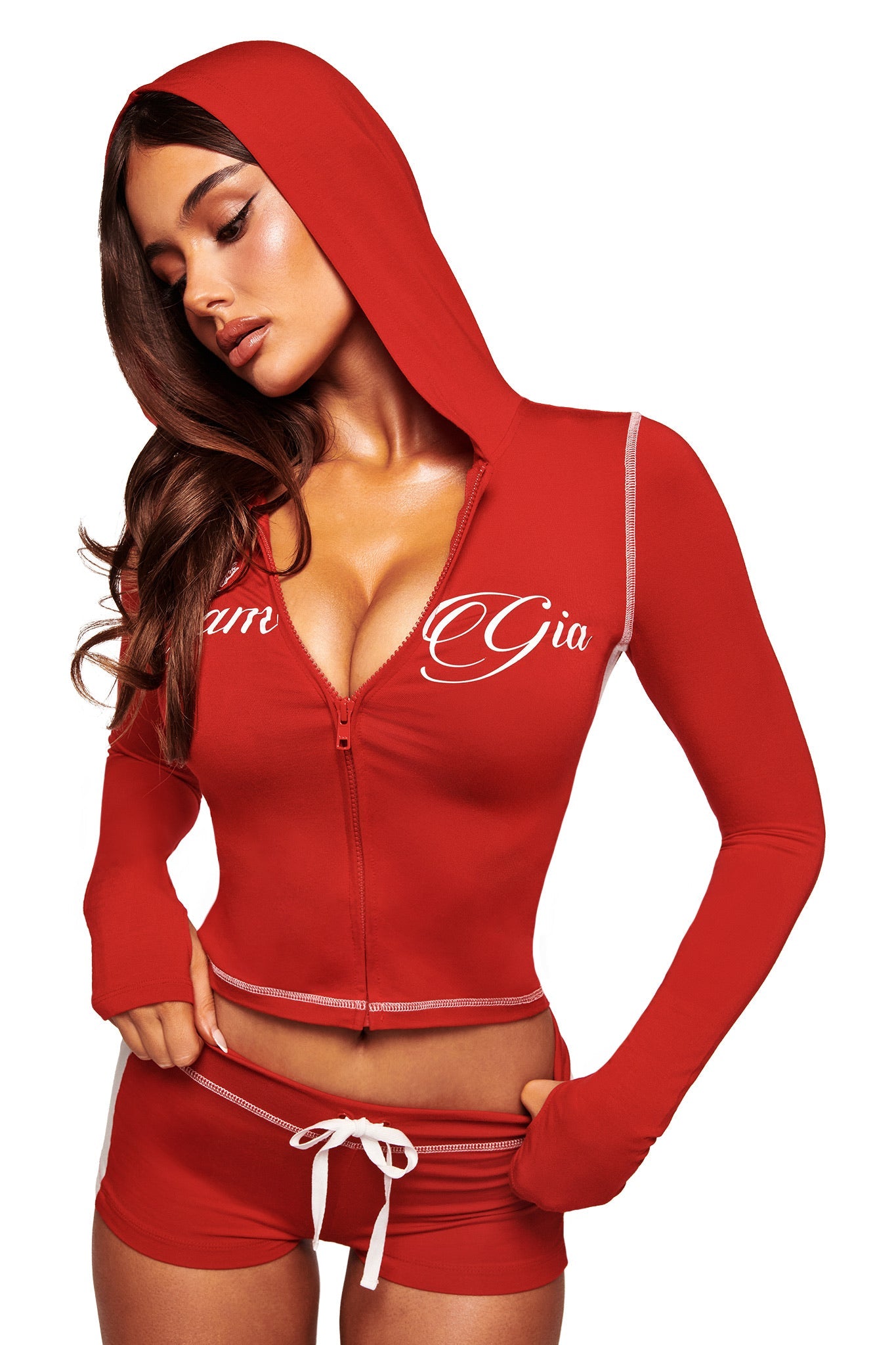 A woman poses against a white background wearing I.AM.GIA's red BLARE BAMBOO PANELLED SHORT with a white drawstring, paired with a matching red hooded zip-up crop top featuring "Gia" on the front.