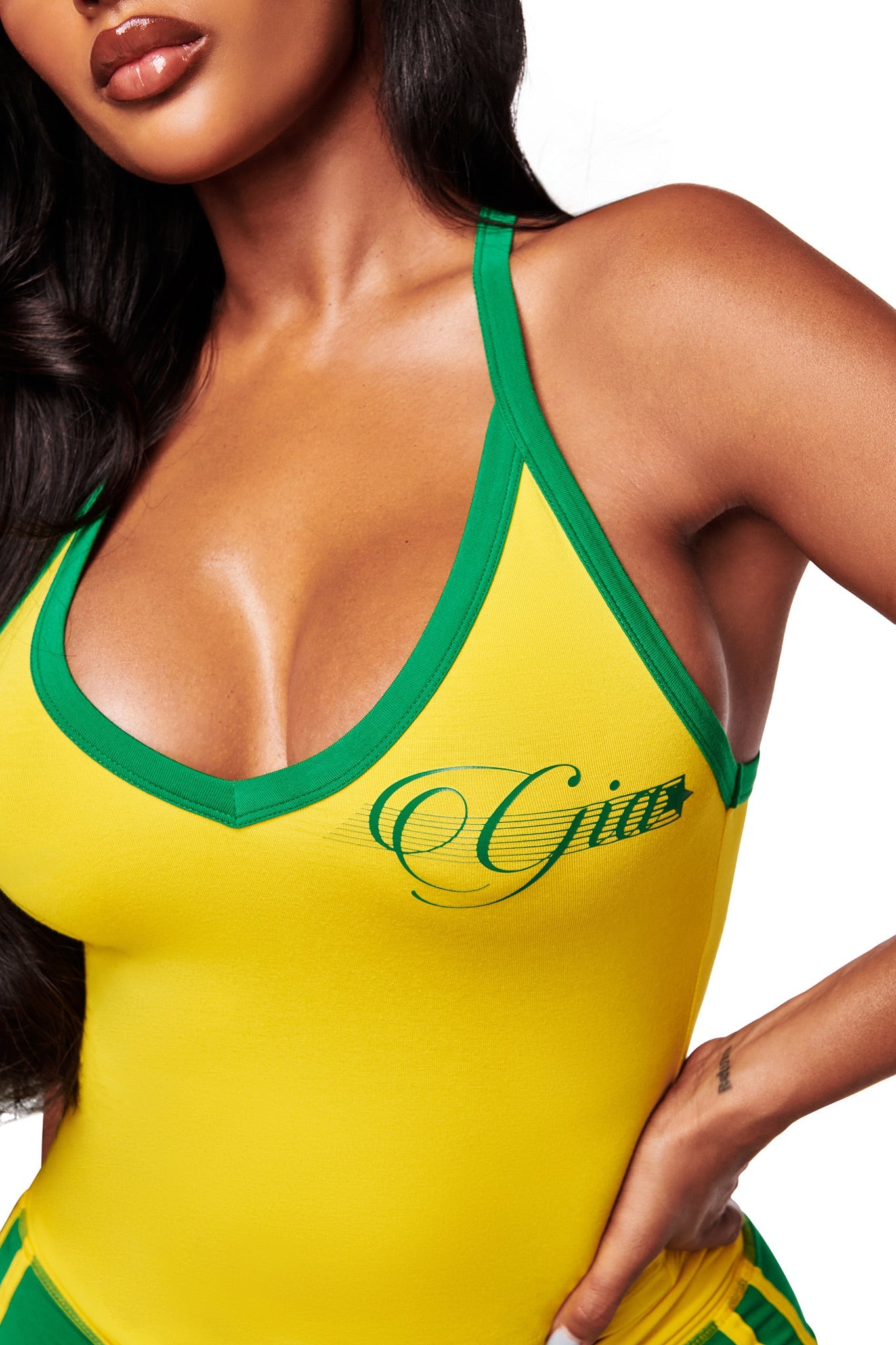 A woman models the I.AM.GIA BLARE V-NECK TOP AND HEADBAND in yellow, featuring green trim and "Gita" script. The cropped image highlights its buttery-soft fabric as she rests her hand on her hip.