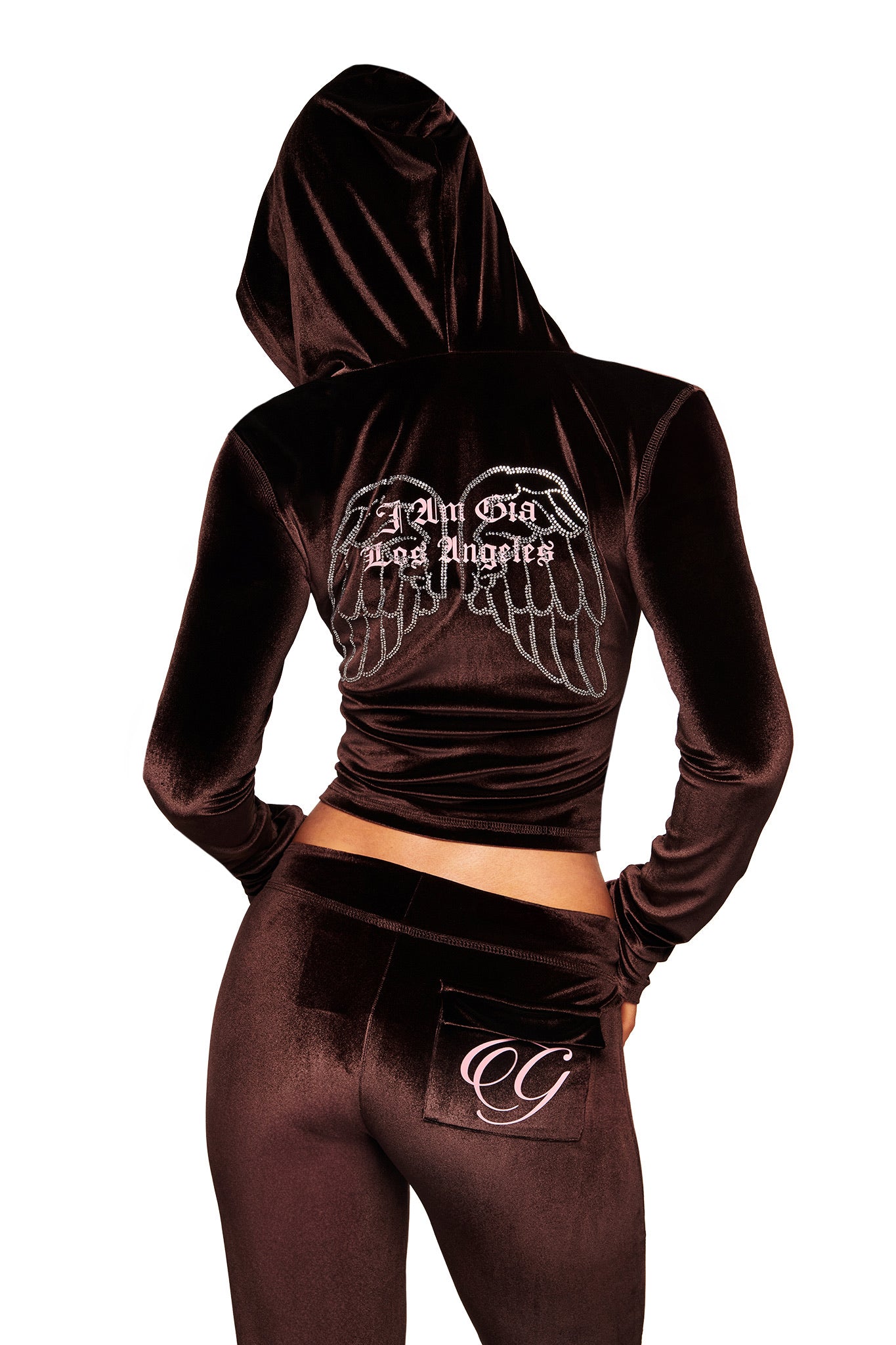 A person wears a dark brown velour tracksuit. The back of the jacket has wing graphics and “Am Glad Los Angeles.” The I.AM.GIA BLARE VELOUR POCKET TRACKPANT in Chocolate features a pocket with a stylized “G” logo.