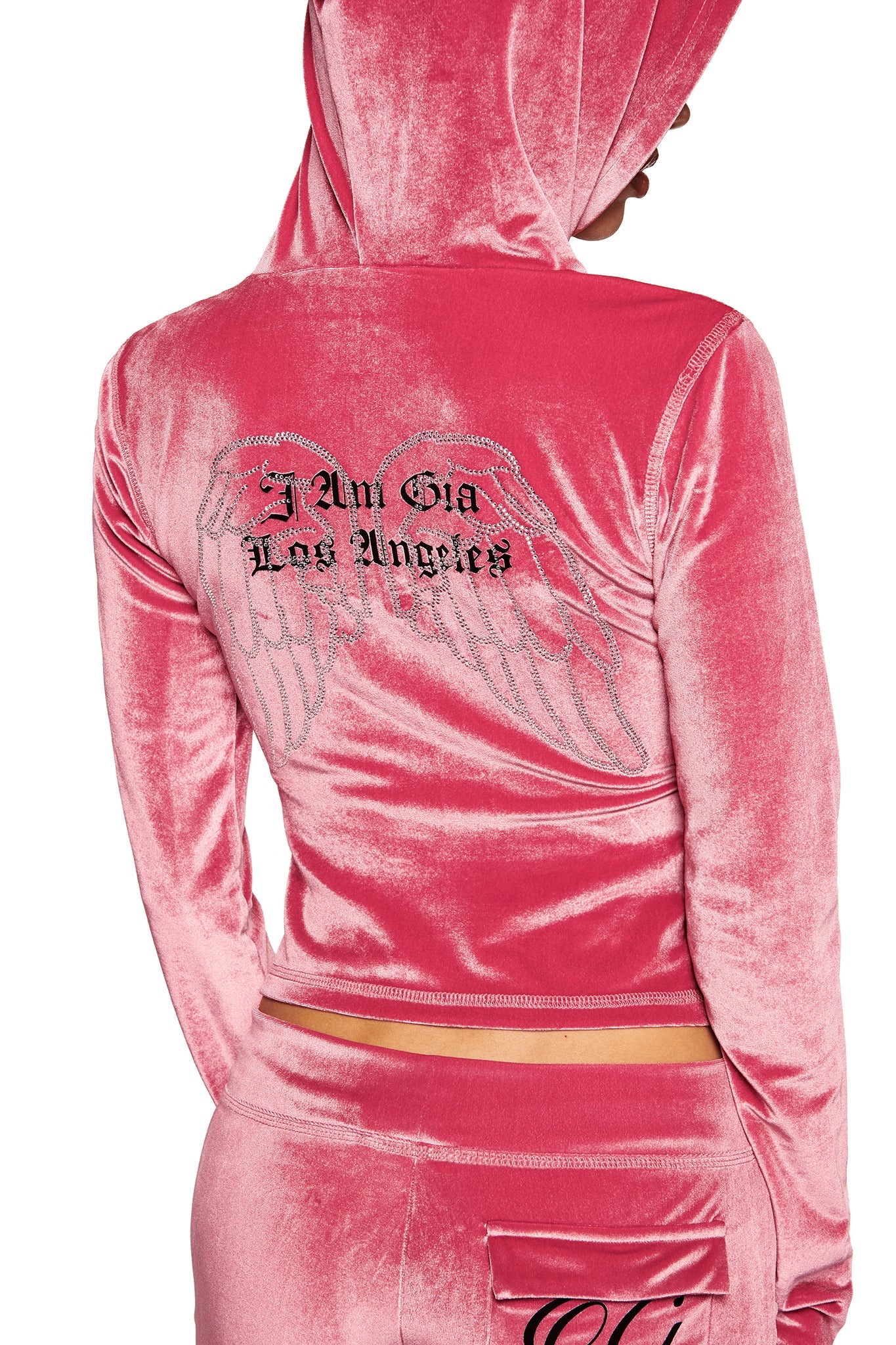 A person wears the I.AM.GIA BLARE VELOUR POCKET HOODIE in pink, featuring diamanté angel wings and “I Am Gia Los Angeles” on the back. The hood is up, showing only the back and part of their face.