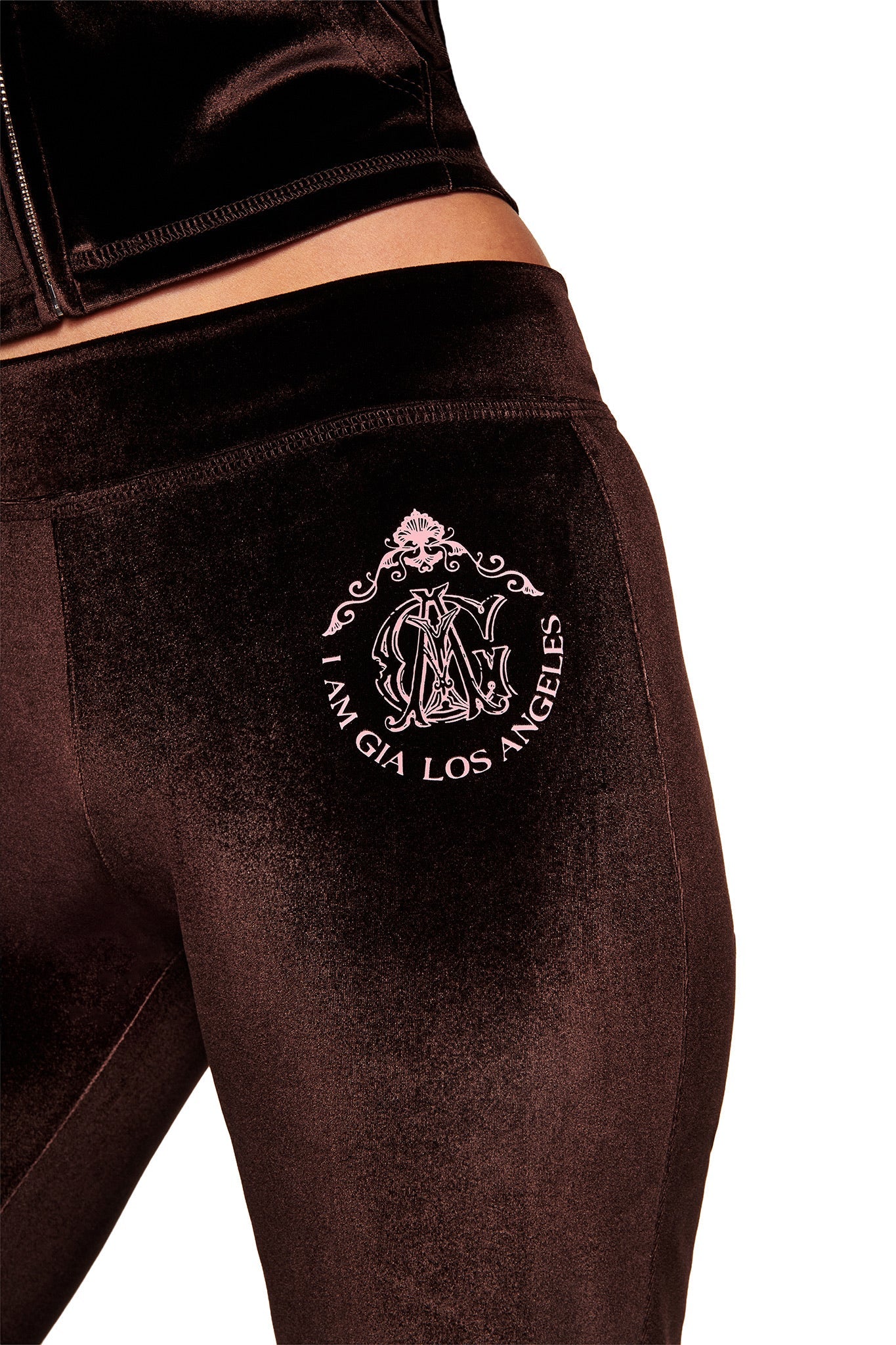 A close-up shows a person wearing I.AM.GIA’s BLARE VELOUR POCKET TRACKPANT in chocolate, featuring a pink crest and "I AM GIA LOS ANGELES" on the back pocket against a white background.