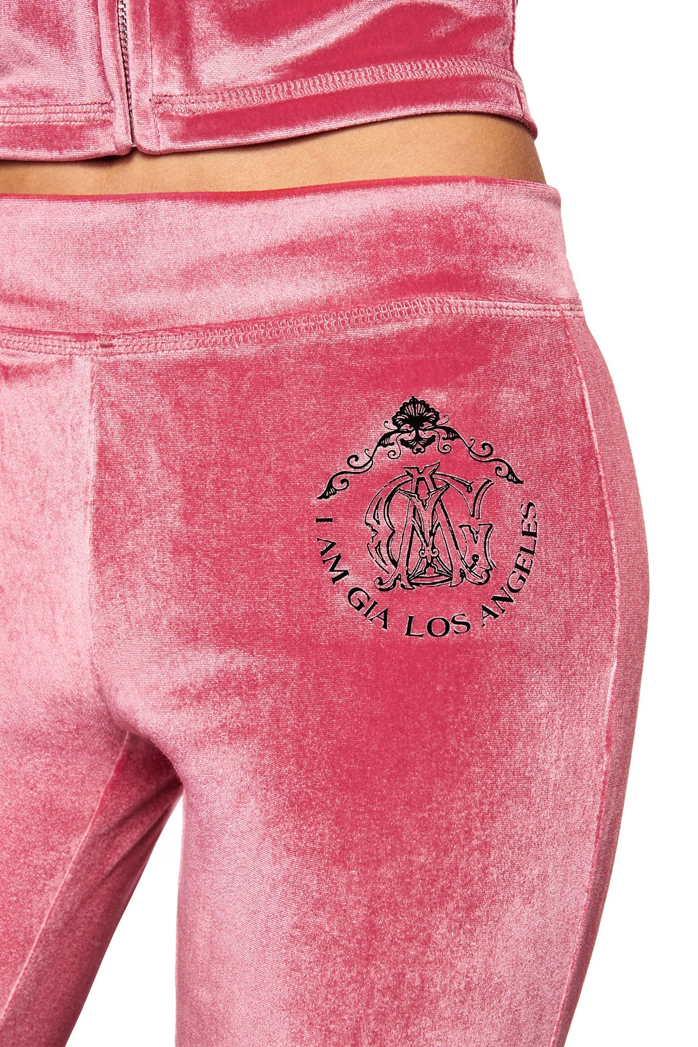 A close-up of the I.AM.GIA BLARE VELOUR POCKET TRACKPANT in pink, featuring a sculpted fit, black emblem, and "I AM GIA LOS ANGELES" print on the hip. The matching jacket is also partially visible.