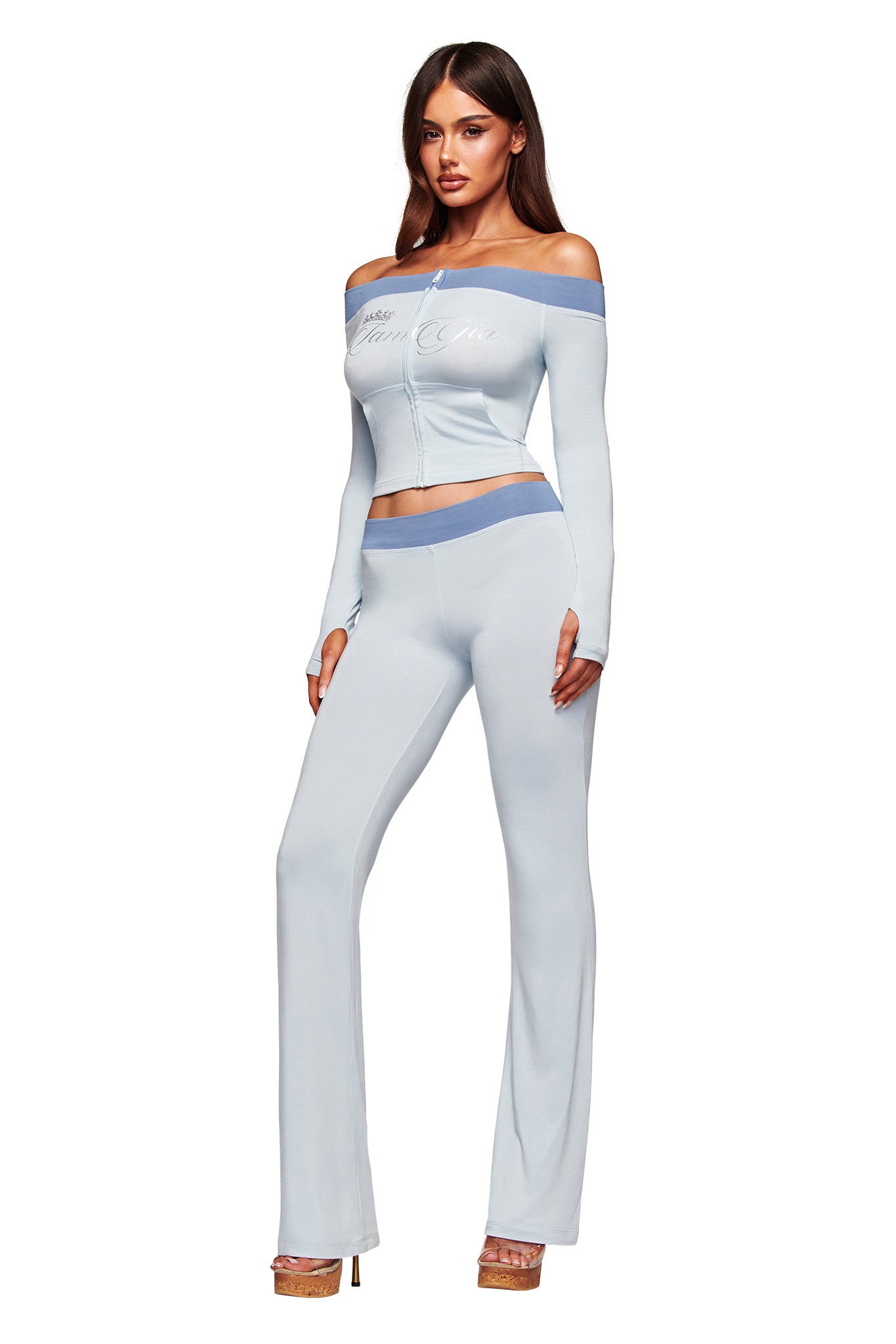 A woman models the I.AM.GIA BLARE OFF SHOULDER TOP - TWO TONE POWDER BLUE with matching flared pants and heeled sandals against a plain white background. The set features buttery-soft viscose fabric and blue trim.