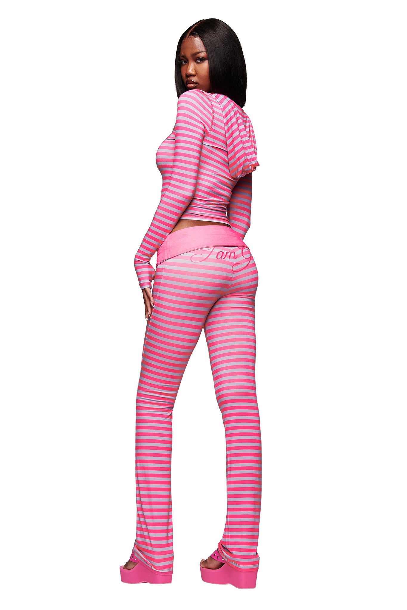 A woman models the I.AM.GIA BLARE FOLDOVER TRACKPANT - HOT PINK STRIPE, featuring light blue stripes, a flared leg, and "I am" on the back, paired with a cropped hoodie as she poses turned away looking over her shoulder.