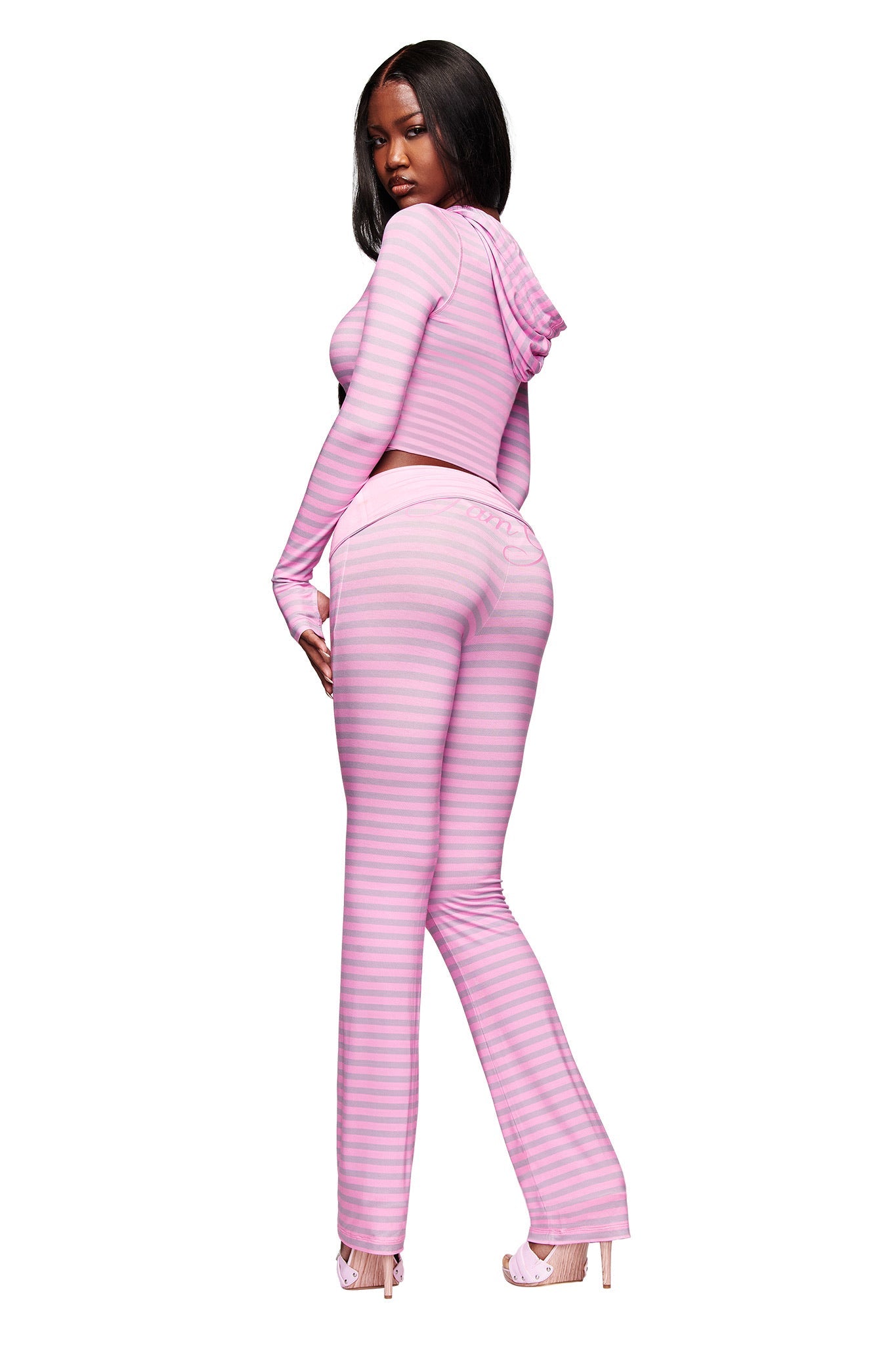 A woman faces backward, glancing over her shoulder. She wears a fitted, long-sleeve hooded crop top and I.AM.GIA BLARE FOLDOVER TRACKPANT – LIGHT PINK STRIPE with a flared leg and high heels, set against a plain white background.