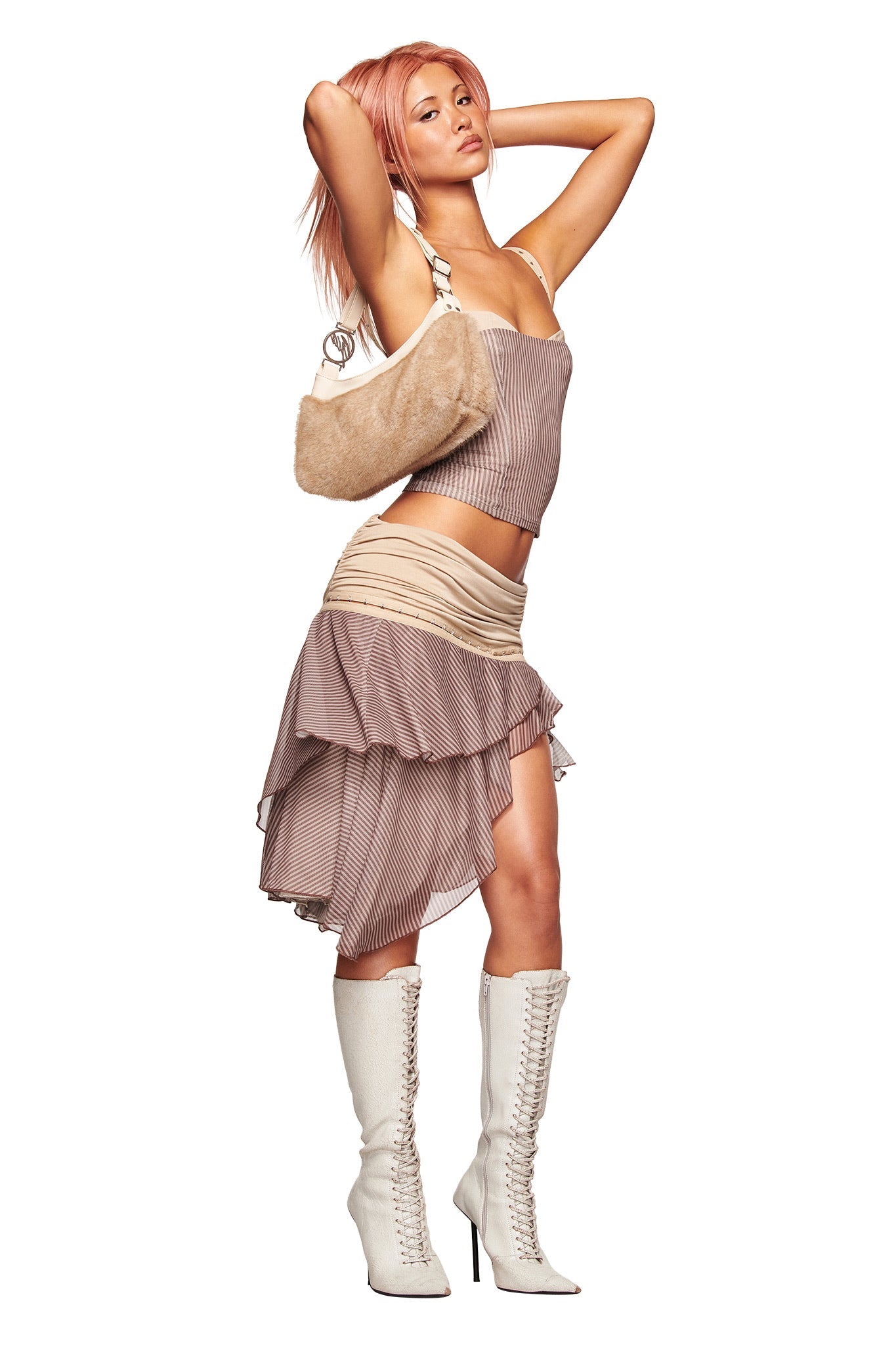 A woman with pink hair poses with her hands behind her head, wearing the I.AM.GIA SEPHI MIDI SKIRT in Mushroom Stripe, a beige sleeveless top, white lace-up knee-high boots, and a tan furry purse against a white background.