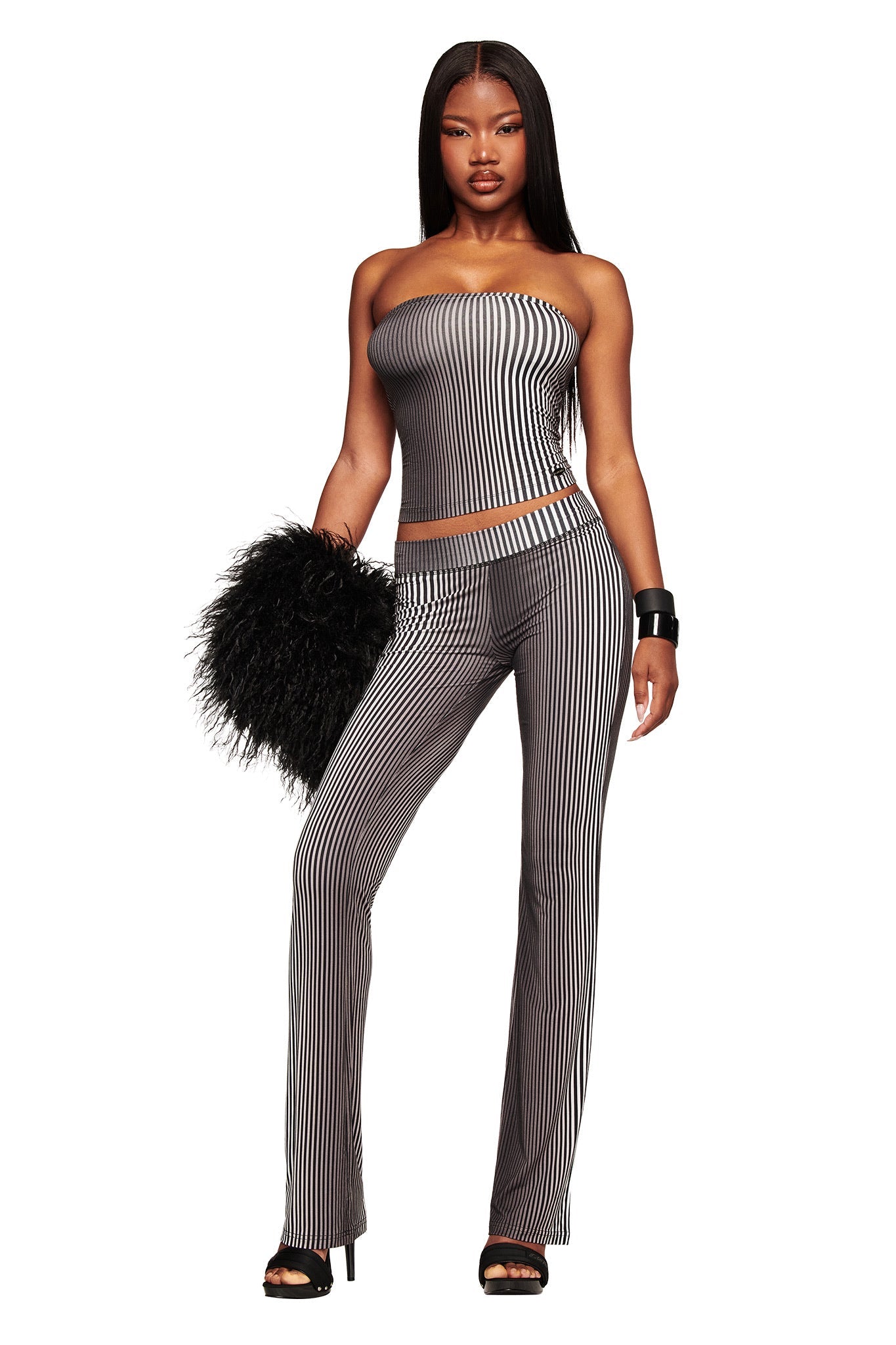 A woman stands confidently in I.AM.GIA’s TRINITY 2-IN-1 SKIRT PANT - STRIPE, paired with a strapless black and white top, black fluffy clutch, heels, and a bangle against a white background.