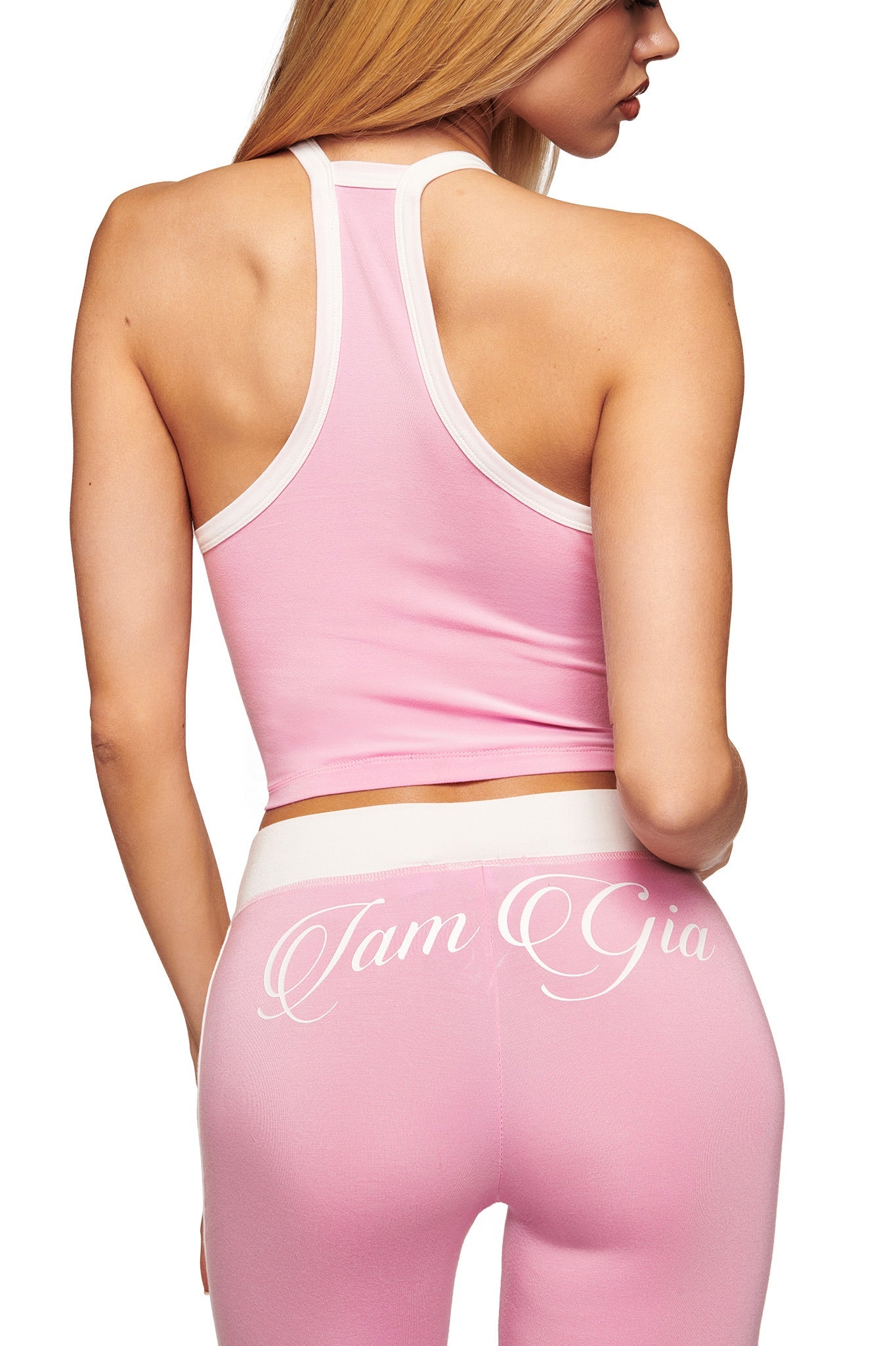 A woman with long blonde hair wears I.AM.GIA’s BLARE PIPING TRACKPANT in baby pink and white, turned away to show off the sporty, high-waisted style and part of her profile.