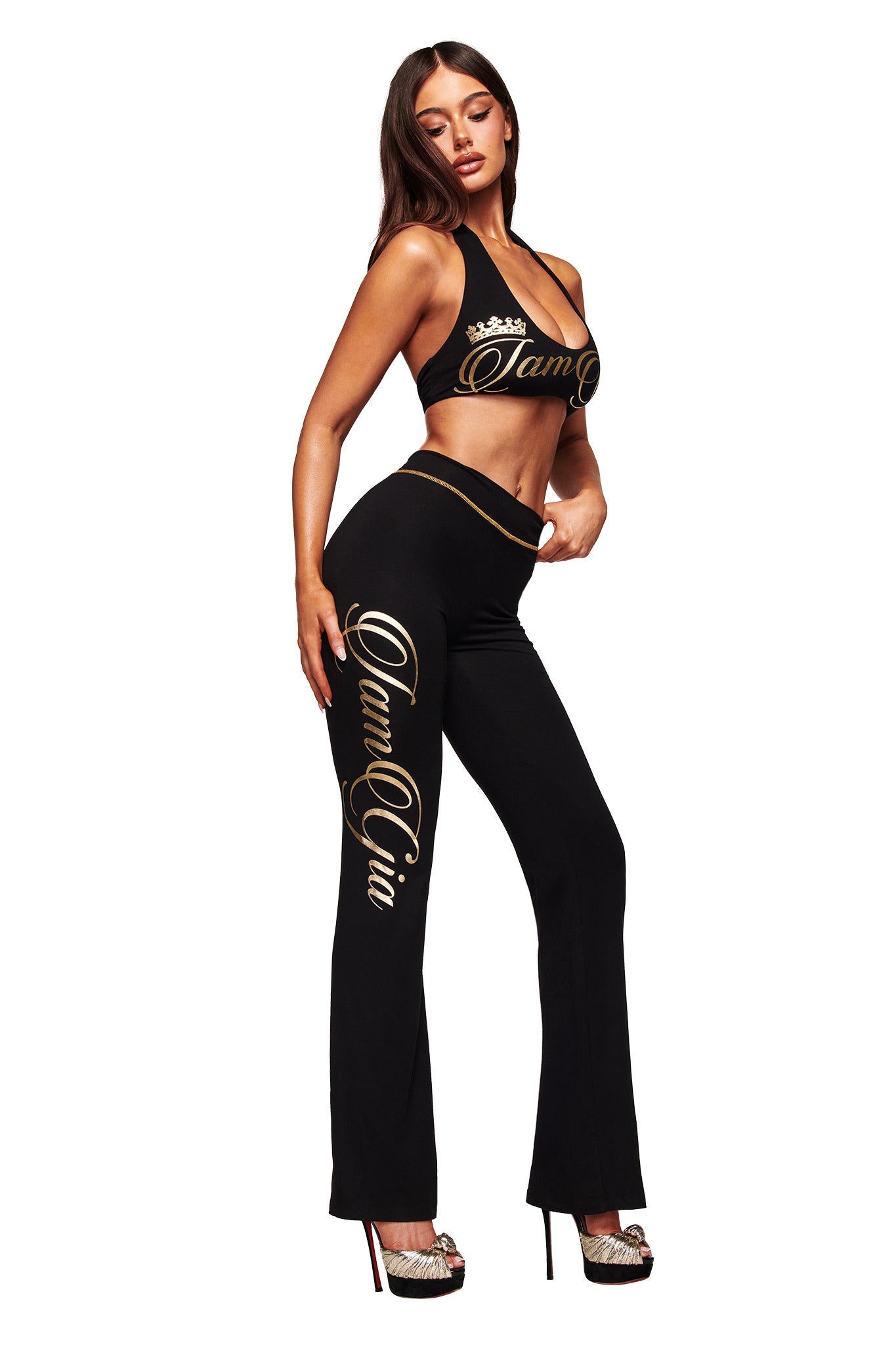 A woman models the I.AM.GIA BLARE FORM HALTER CROP TOP in black and gold, paired with high heels, posing against a white background.