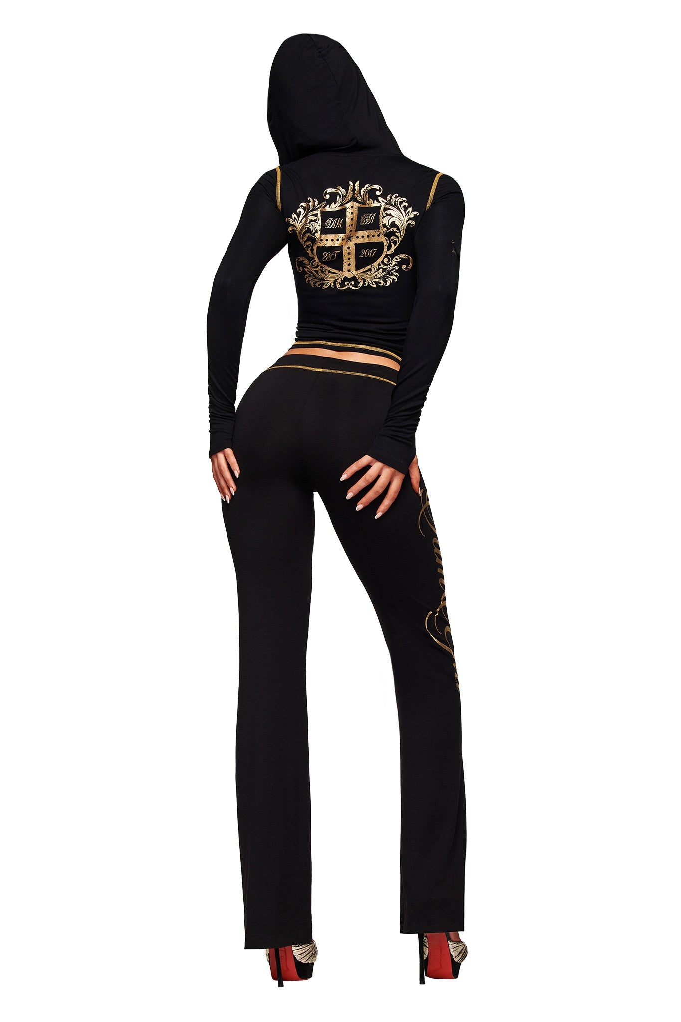 A person in I.AM.GIA's BLARE FORM TRACKPANT in black and gold, featuring a fitted design with metallic print and ornate details, poses with their back to the camera, hands on hips, wearing red high heels.