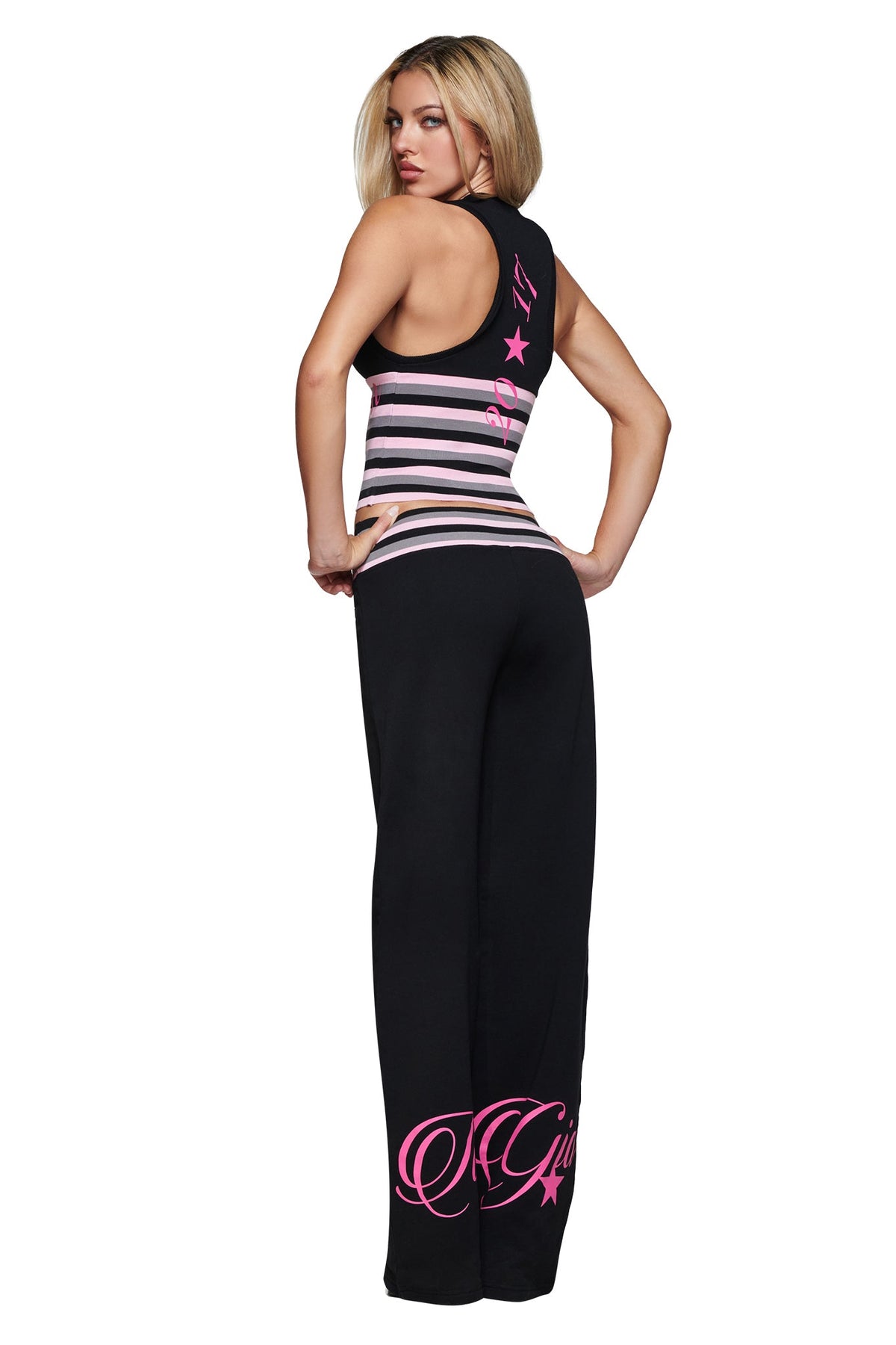 A woman stands with her back turned, wearing the I.AM.GIA CANDI TOP in black—a V-neck racerback style featuring pink and white horizontal stripes—paired with black bodycon pants accented by pink script and star designs near the ankle and waist.