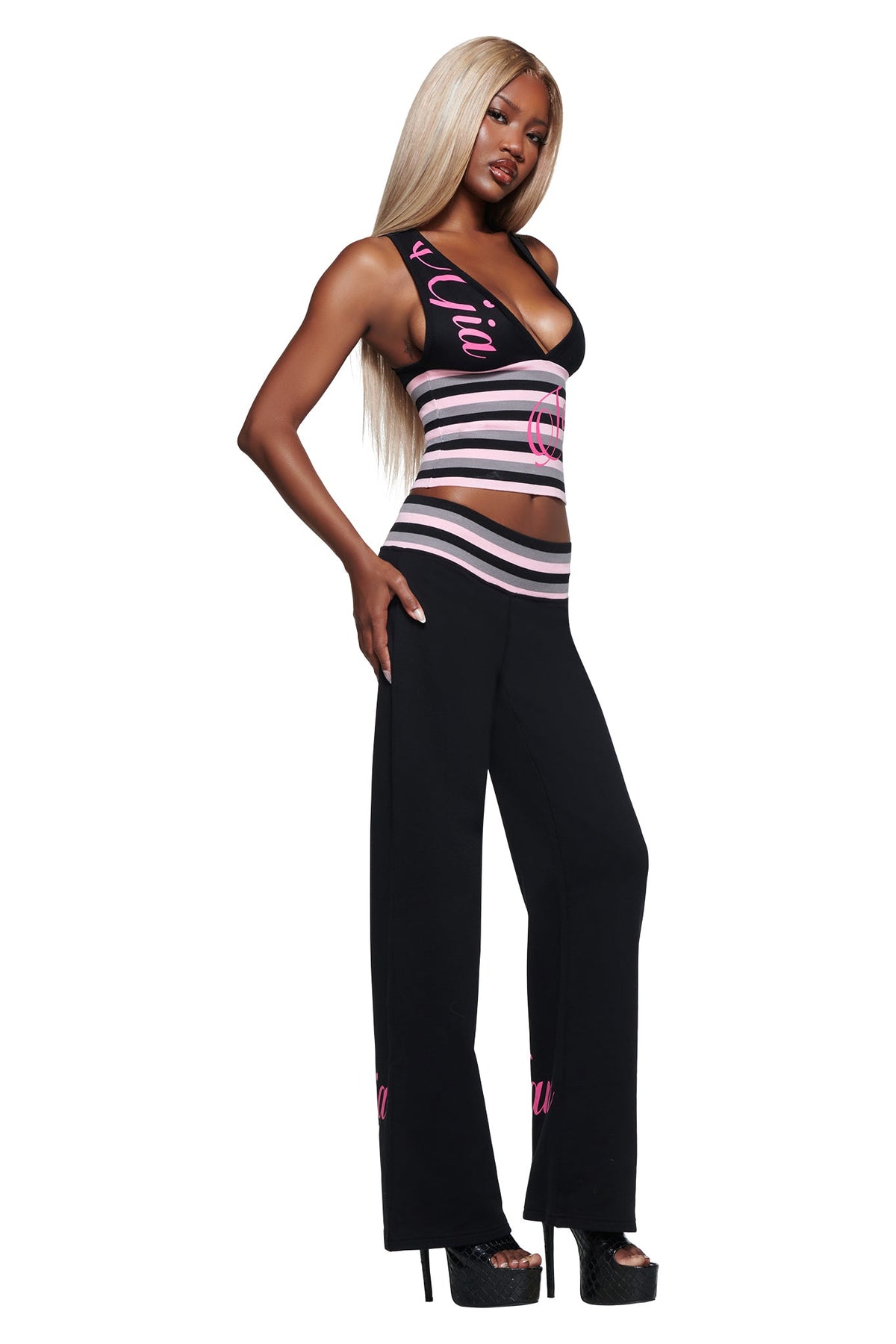 A woman with long blond hair wears the I.AM.GIA CANDI TOP in black, featuring a V-neck racerback design with pink and white stripes, paired with matching bodycon black pants and platform heels, standing confidently.